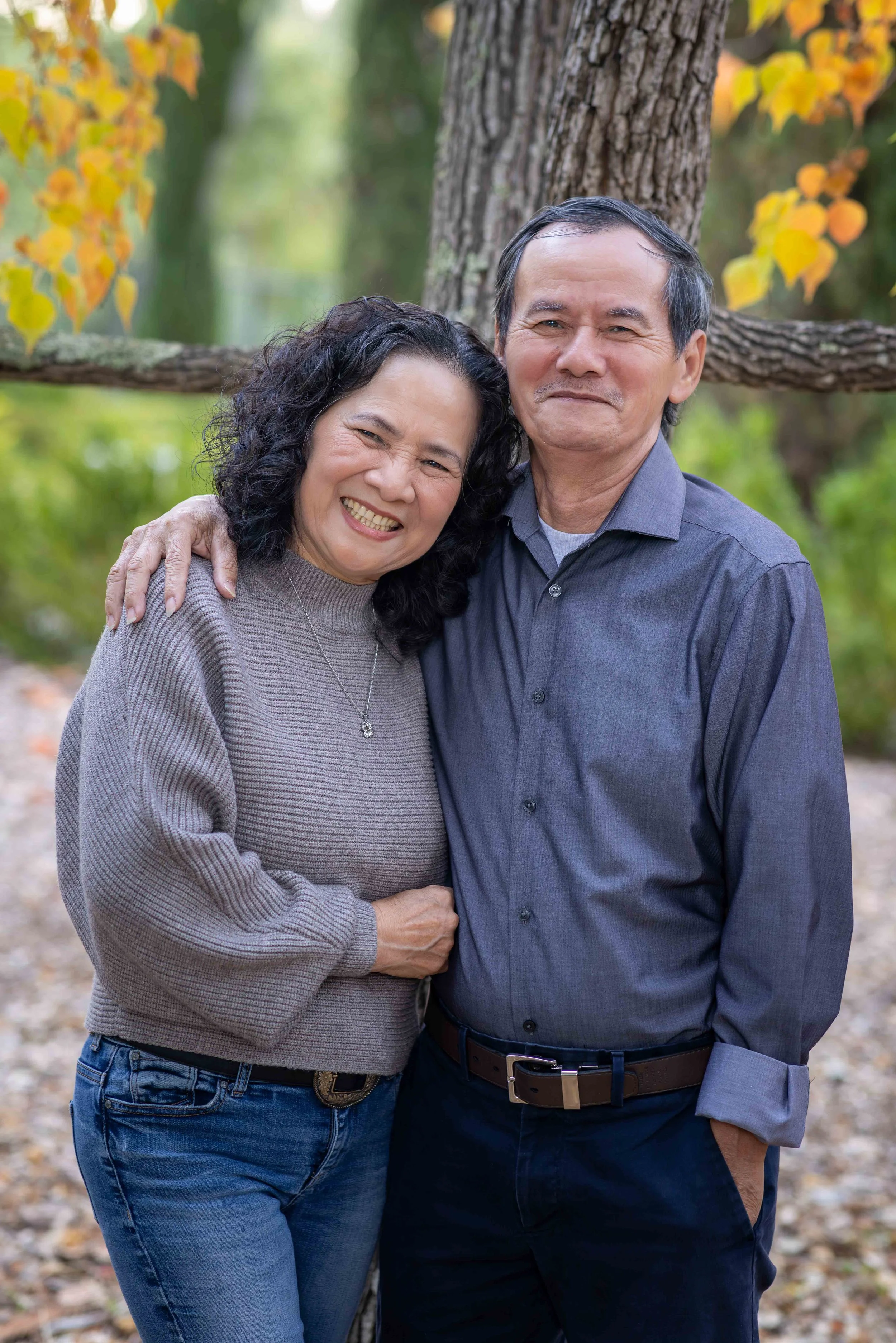 A smiling middle-aged woman and man embracing outdoors in front of a large tree with fall foliage.