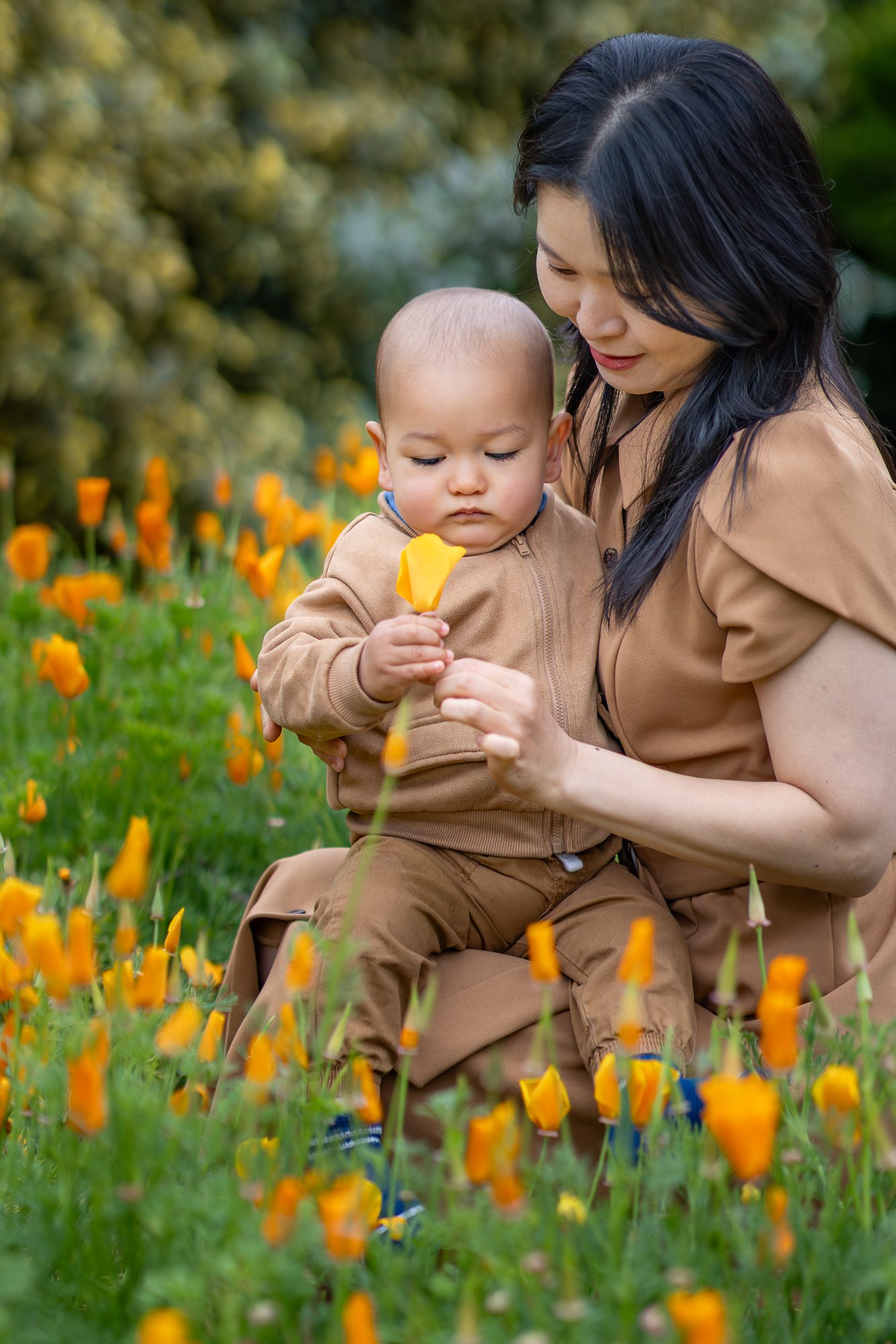 A woman and a young child sitting among orange flowers in a garden. The woman is looking down at the child, who is holding a yellow flower and examining it.