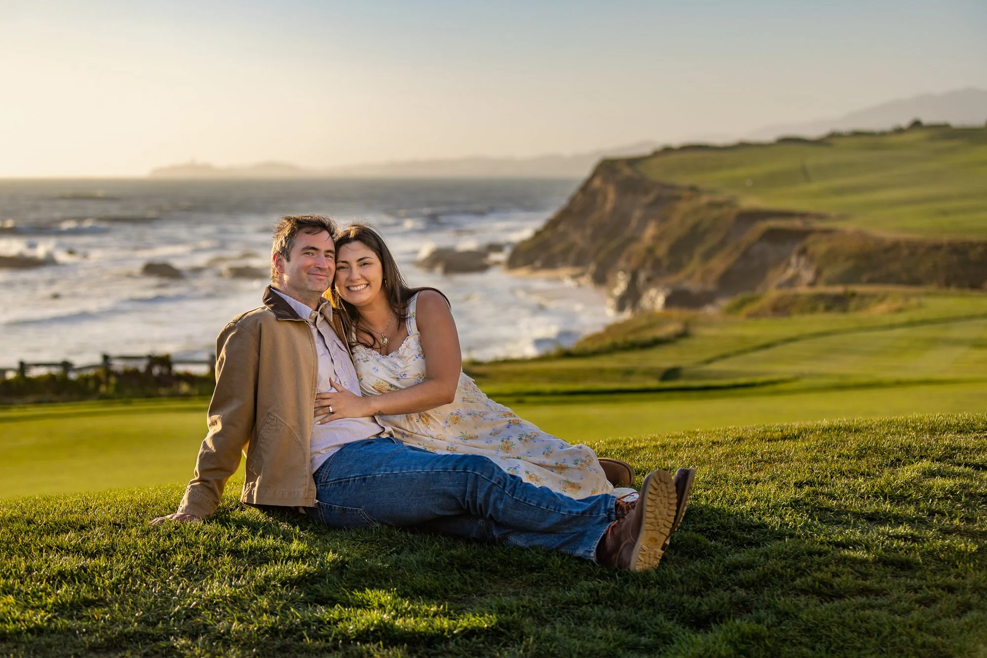 A couple sitting on a grassy area outdoors near a coastline with cliffs and ocean, smiling and enjoying the sunset.