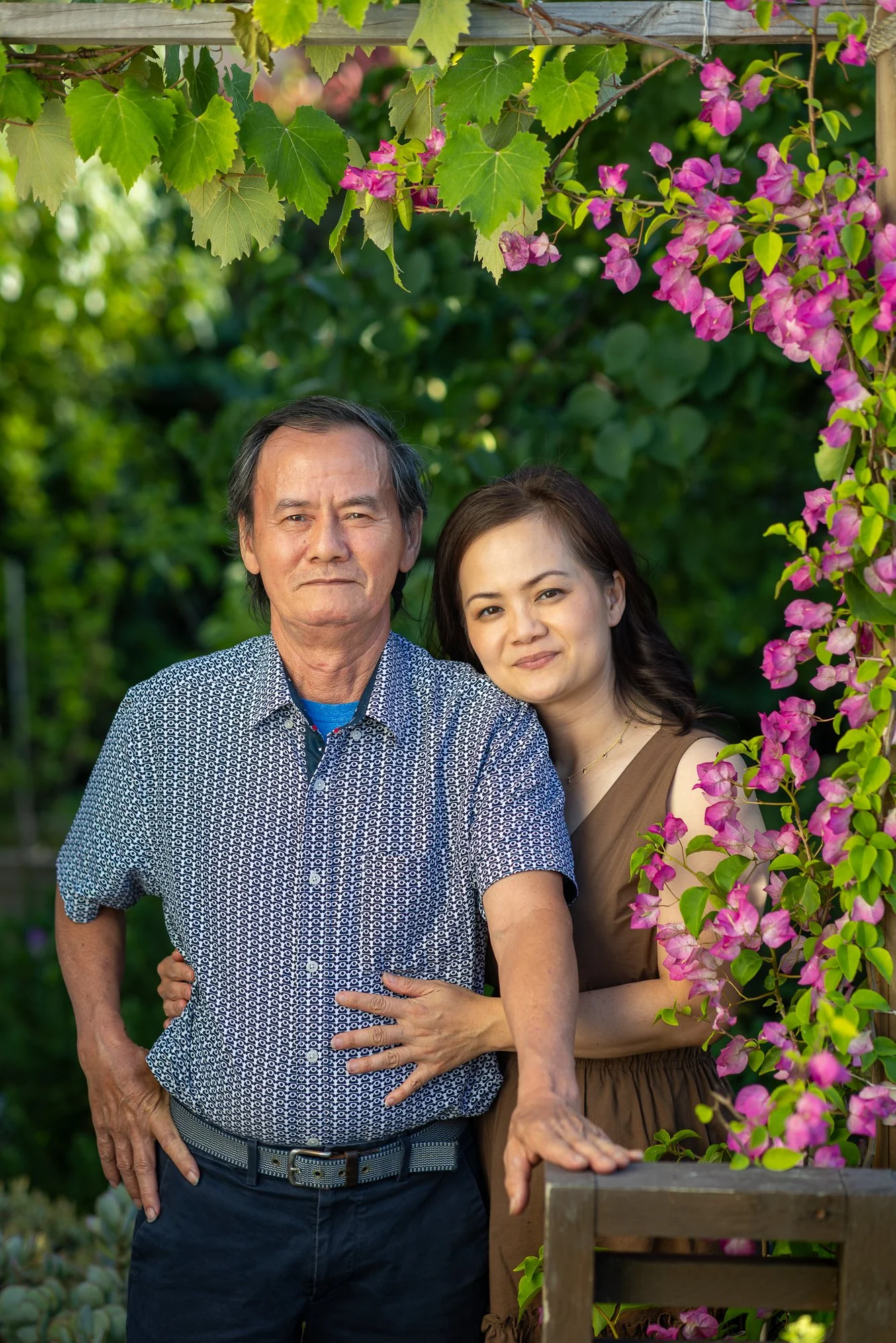 An older man and a young woman standing together outdoors with lush green background and pink flowers, posing for a photo.