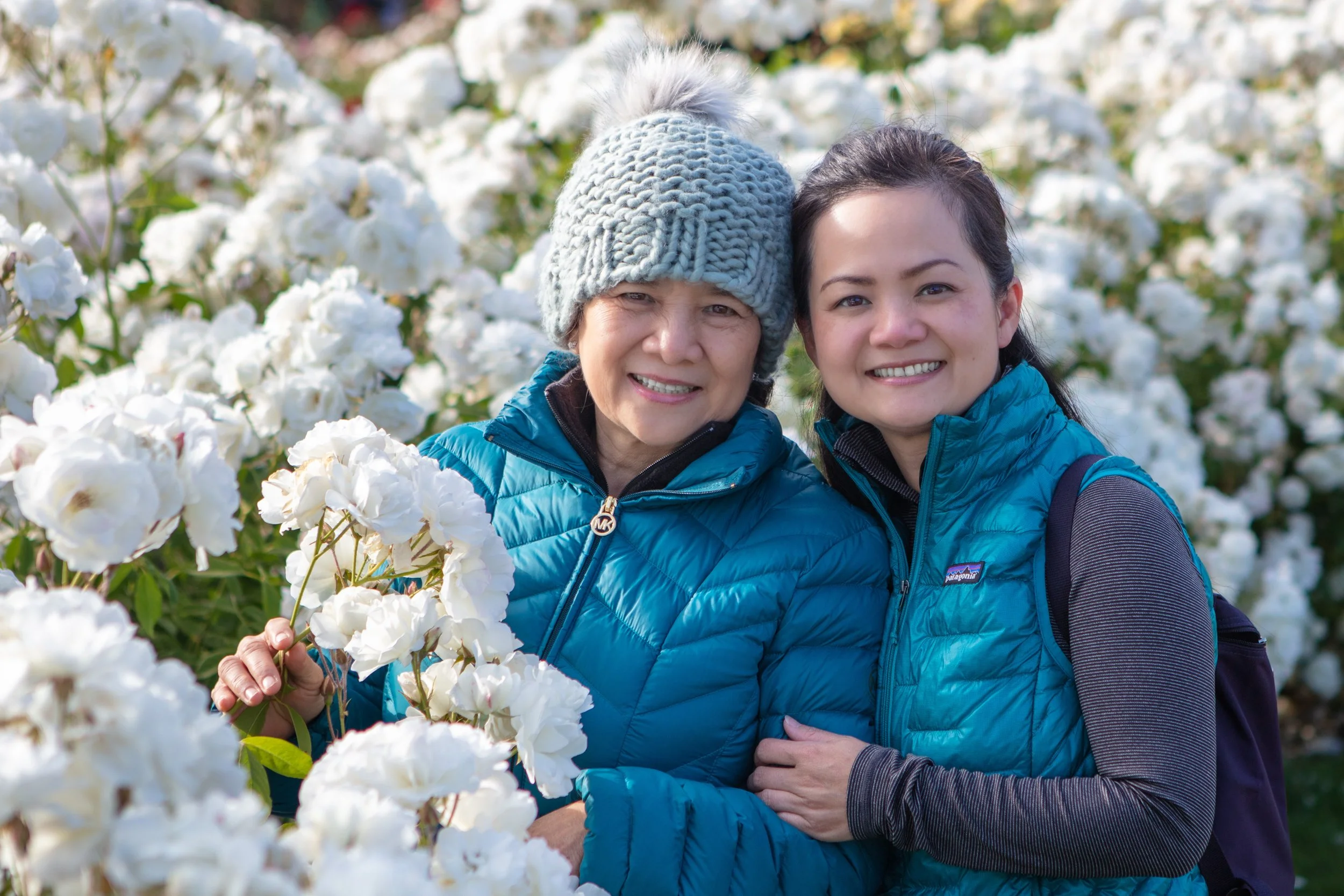 Two women smiling, standing close together outdoors among white flowering bushes, wearing blue jackets and one with a grey knit beanie.