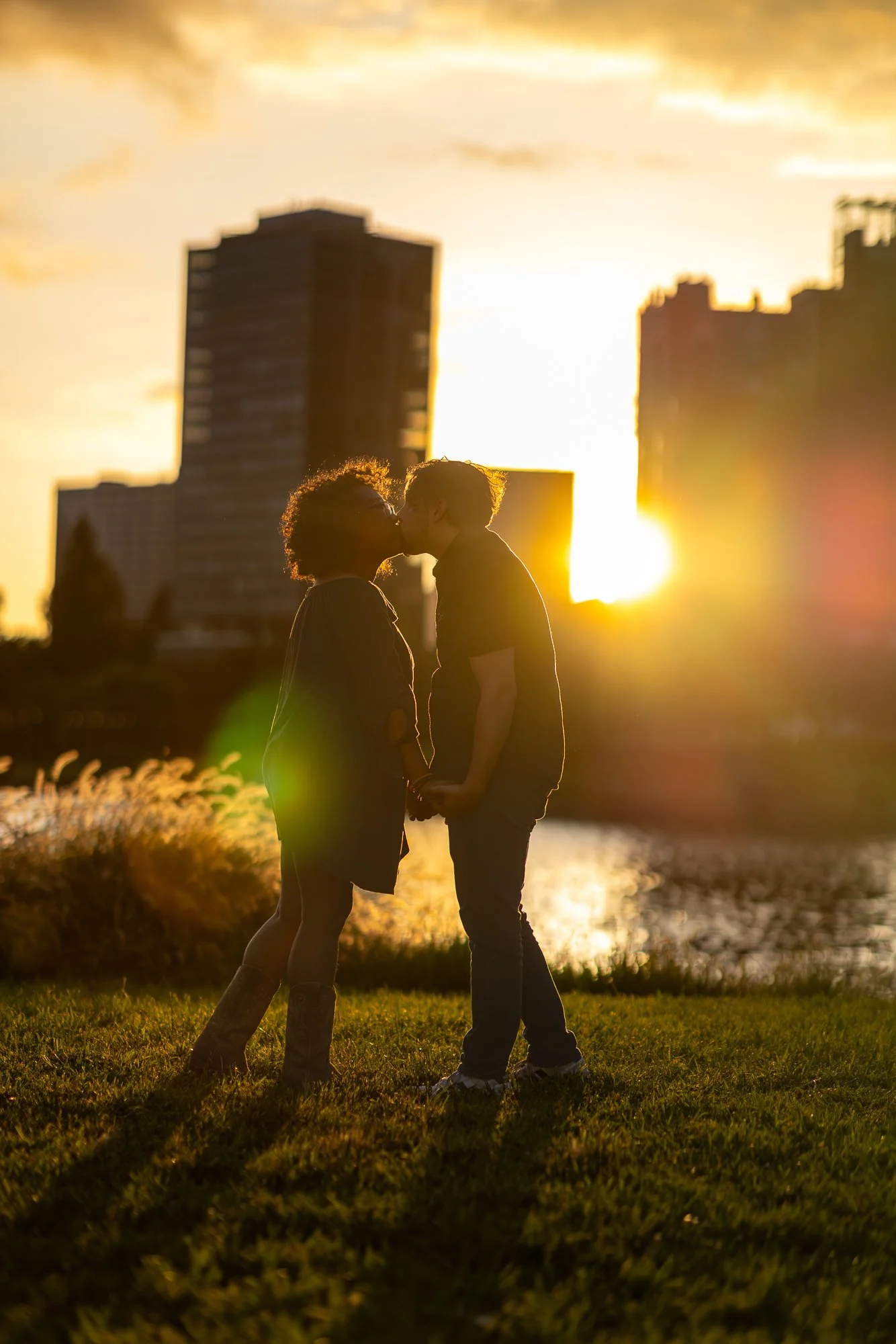 A silhouette of two people, likely a couple, sharing a kiss or intimate moment at sunset by a body of water with city buildings in the background.