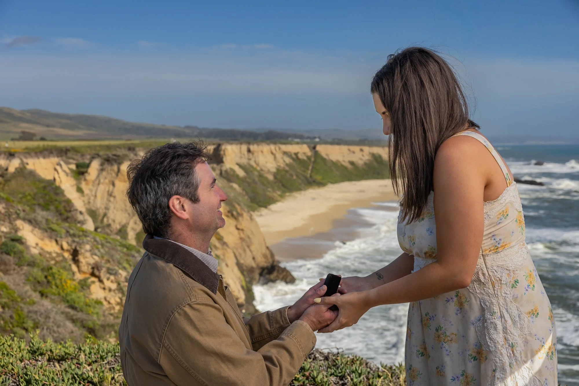 A man proposing to a woman by the ocean on a sunny day, with cliffs and waves in the background.