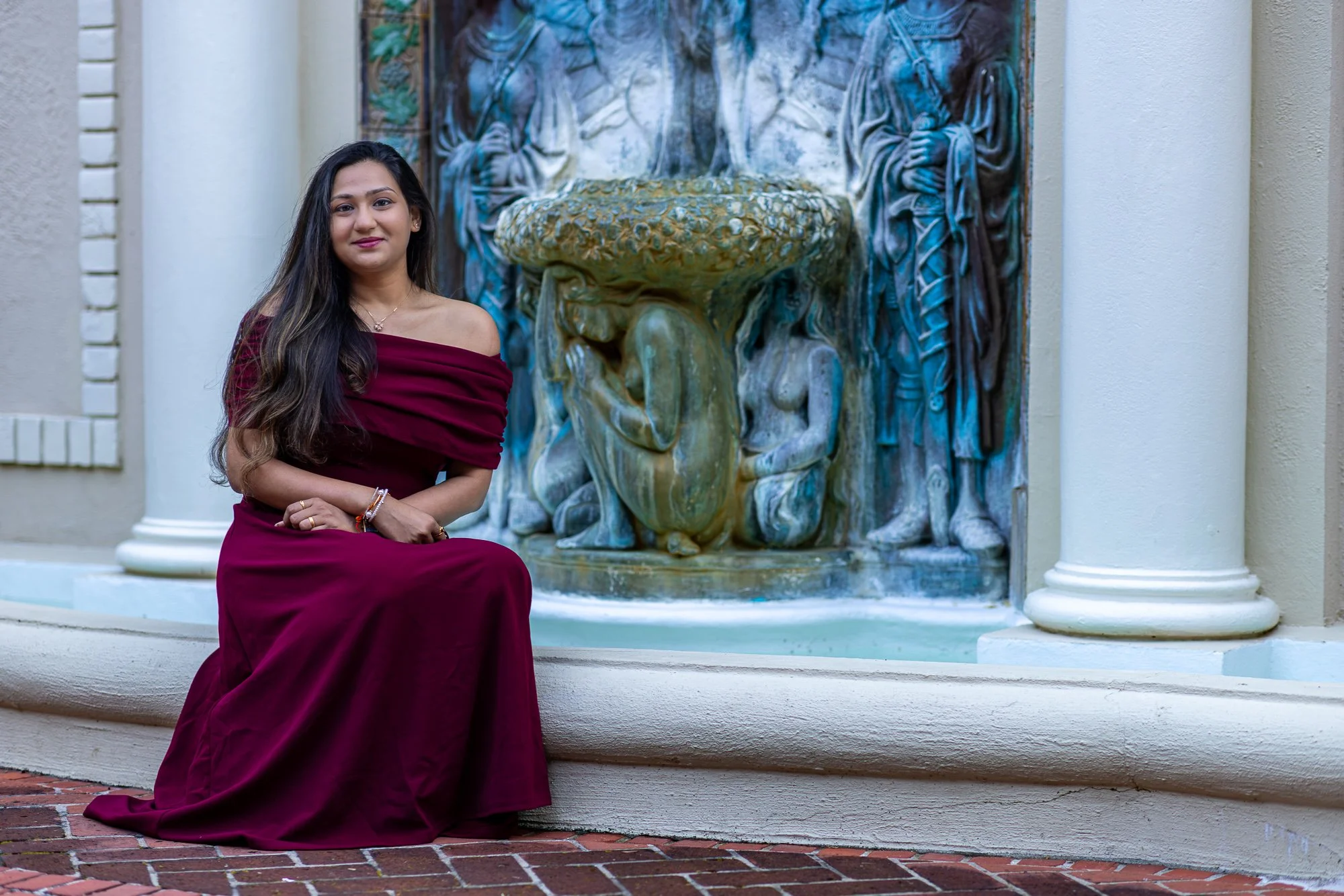 A woman in a strapless burgundy dress sitting on a ledge in front of a decorative water fountain.
