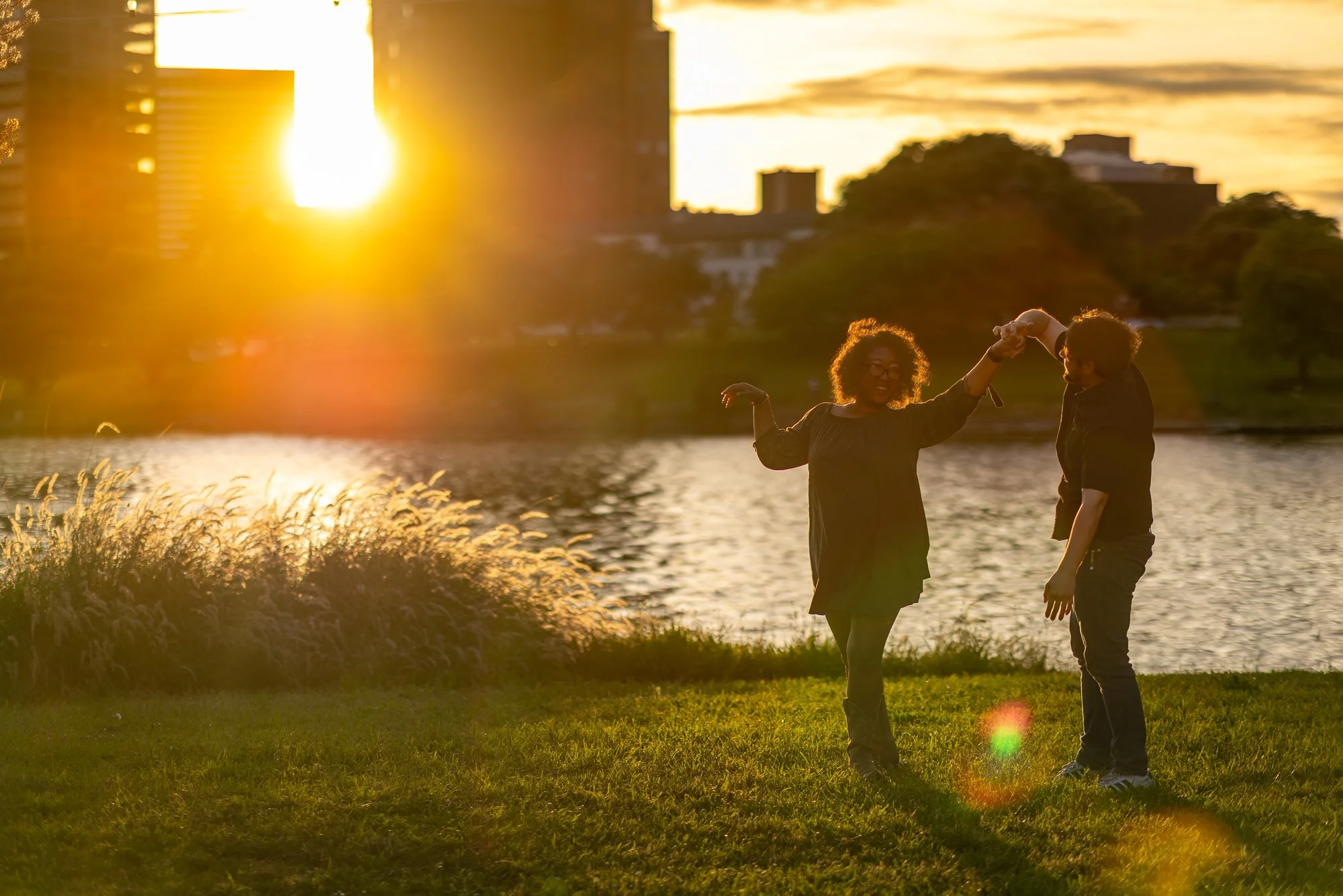 A couple dancing by a river at sunset, with the sun setting behind city buildings in the background.