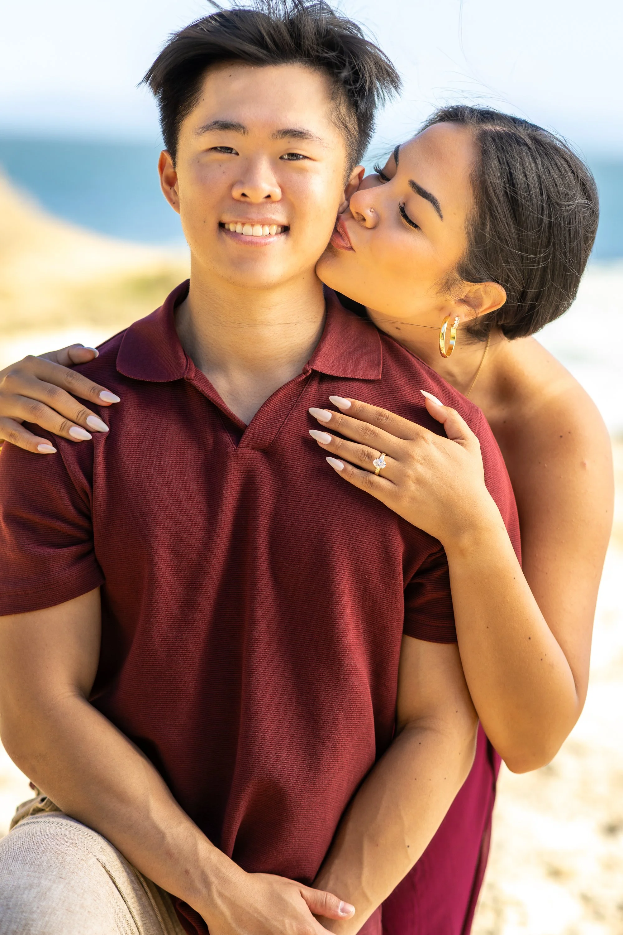 A woman with short dark hair and hoop earrings is kissing a young man on the cheek, both smiling, at the beach.