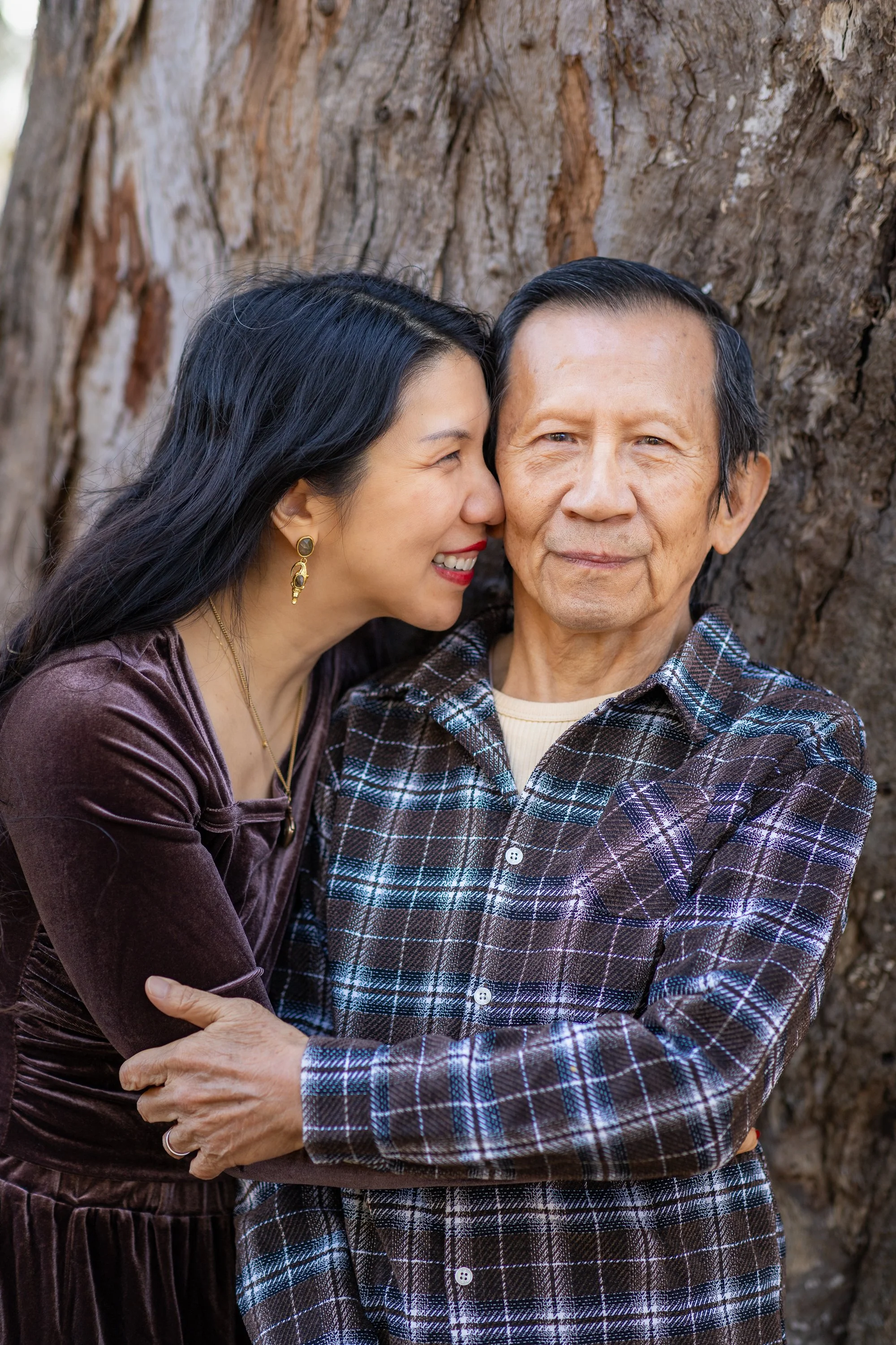 A woman with long black hair leaning in to kiss a smiling elderly man on the cheek, standing outdoors in front of a large tree trunk.