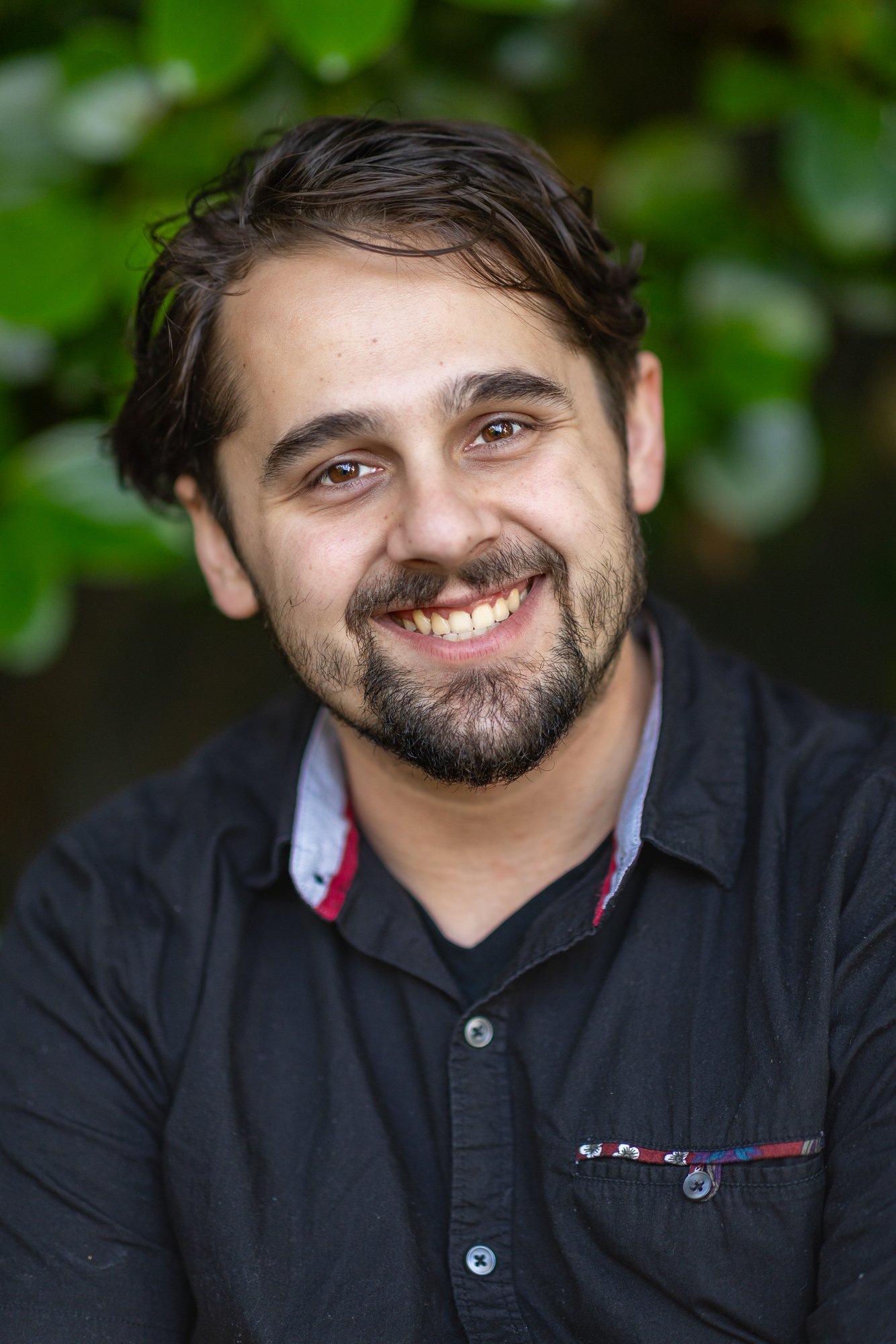 Close-up of a young man smiling, with brown eyes, dark brown hair, and a beard, wearing a black shirt with a patterned pocket, outdoors with green foliage in the background.