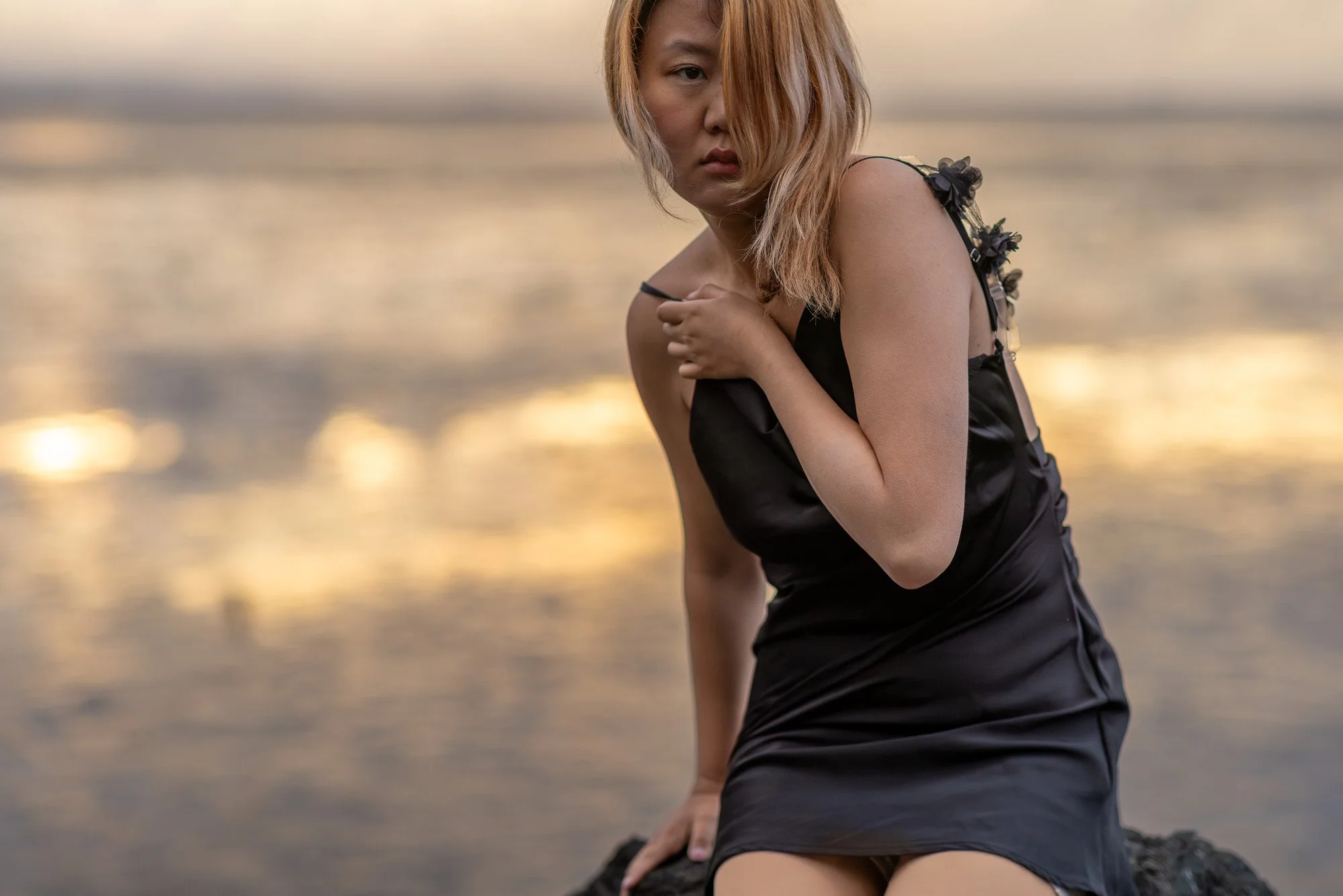 Woman in black silk dress seated on rock at sunset with golden San Francisco Bay reflection behind, Dumbarton Bridge photography Bay Area