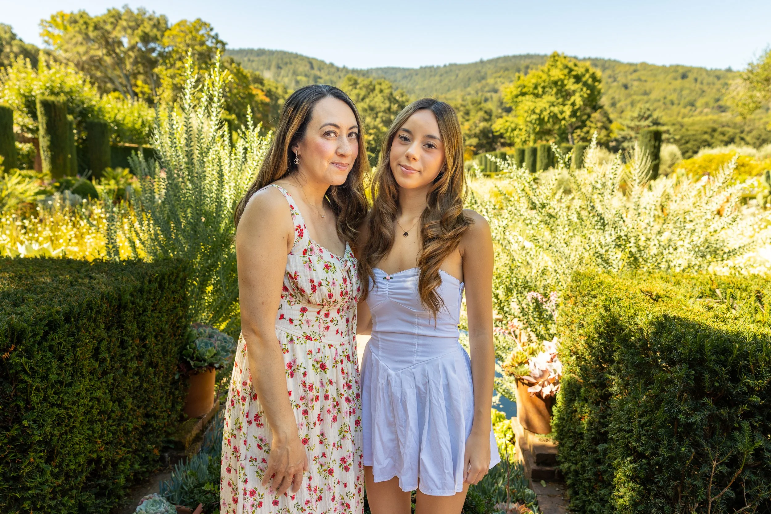 Two women standing together outdoors in a garden with various lush green plants and trees in the background, both smiling gently at the camera.