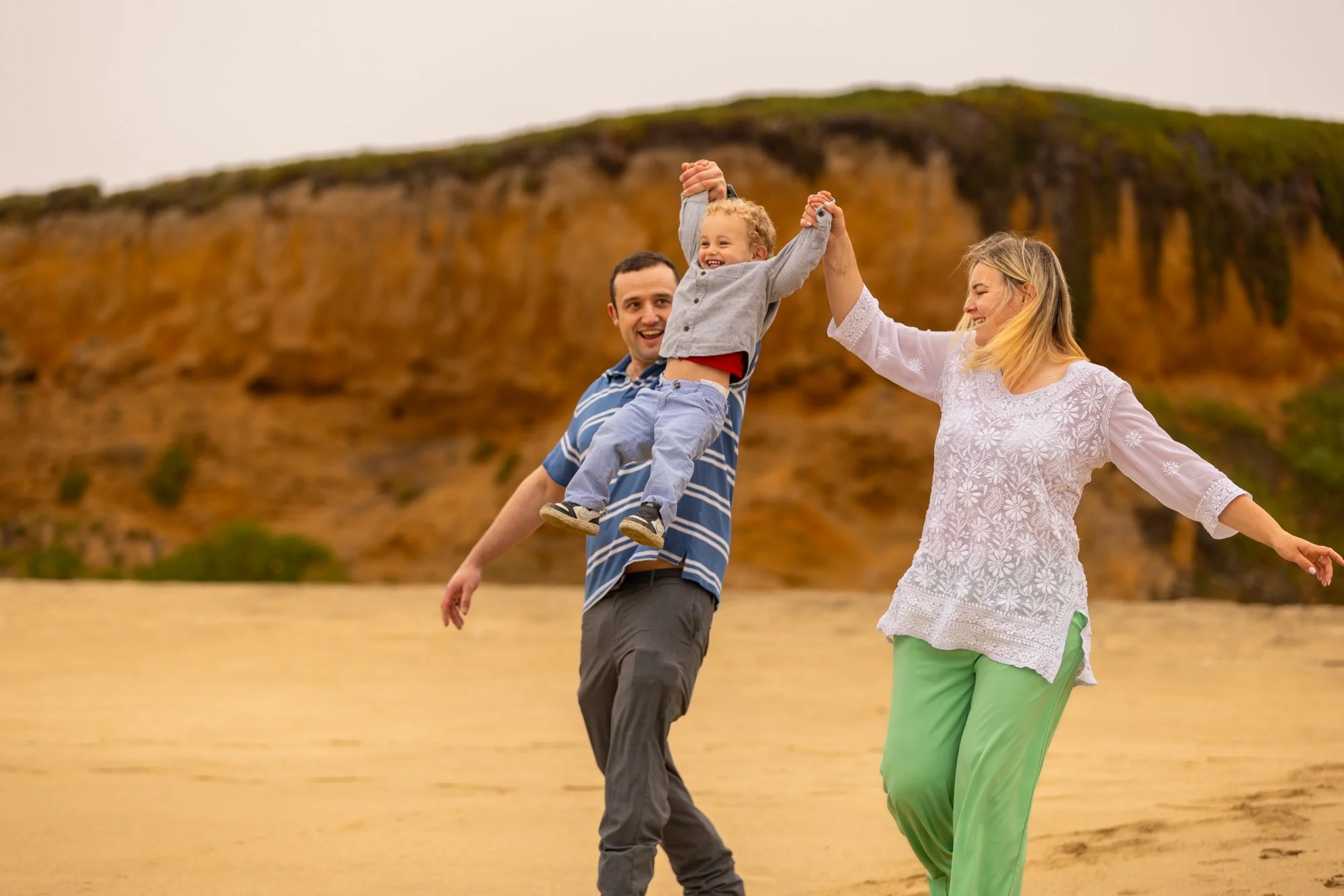 A family of three toddler, father, and mother enjoying a walk outdoors at the beach with rainbow cliffs in the background. The toddler is being held up with arms stretched out by the father, and the mother is smiling and walking close by.