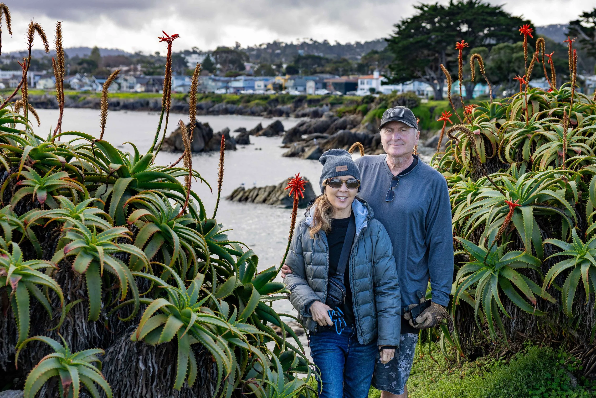 Red aloe flowers framing a couple with Monterey Bay visible across the water — full-day adventure photography session on 17 Mile Drive, Pacific Grove.
