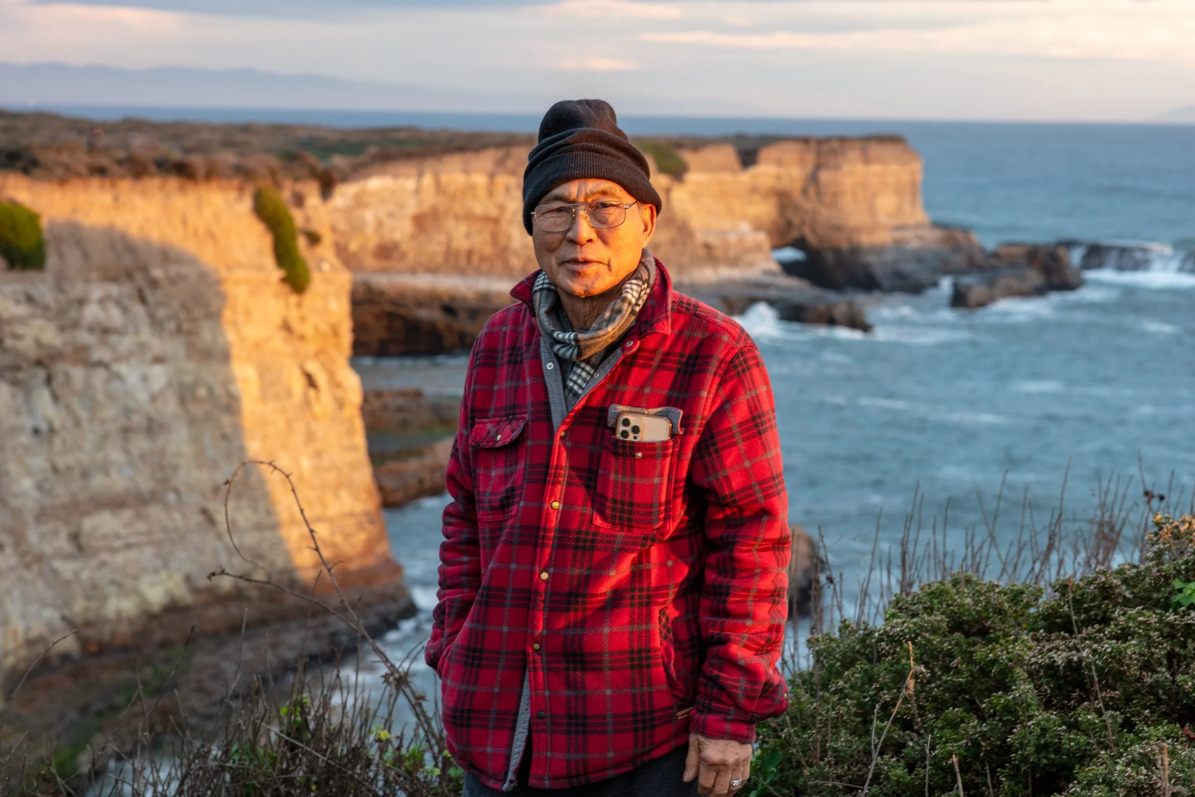 A man wearing glasses, a black beanie, a red and black plaid jacket with a phone in the chest pocket, and a scarf, standing outdoors near cliffs and the ocean during sunset.