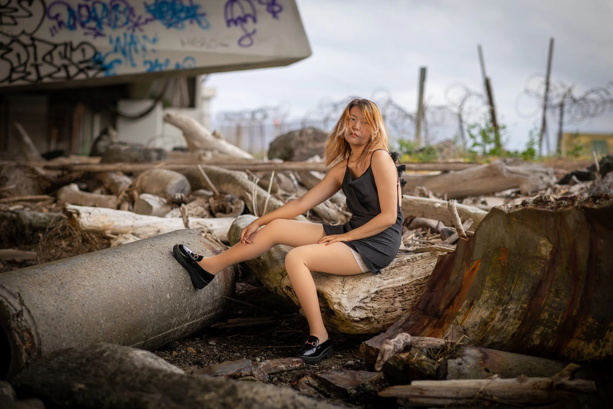 Woman in black dress seated on concrete pipe with razor wire fence background, Dumbarton Bridge shoreline trail editorial portrait Bay Area
