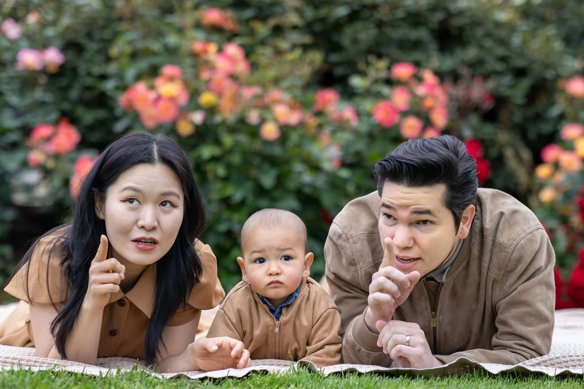 Family of three, including a woman, a man, and a young boy, lying on their elbows outdoors in a garden with blooming flowers in the background, engaging in conversation or pointing.