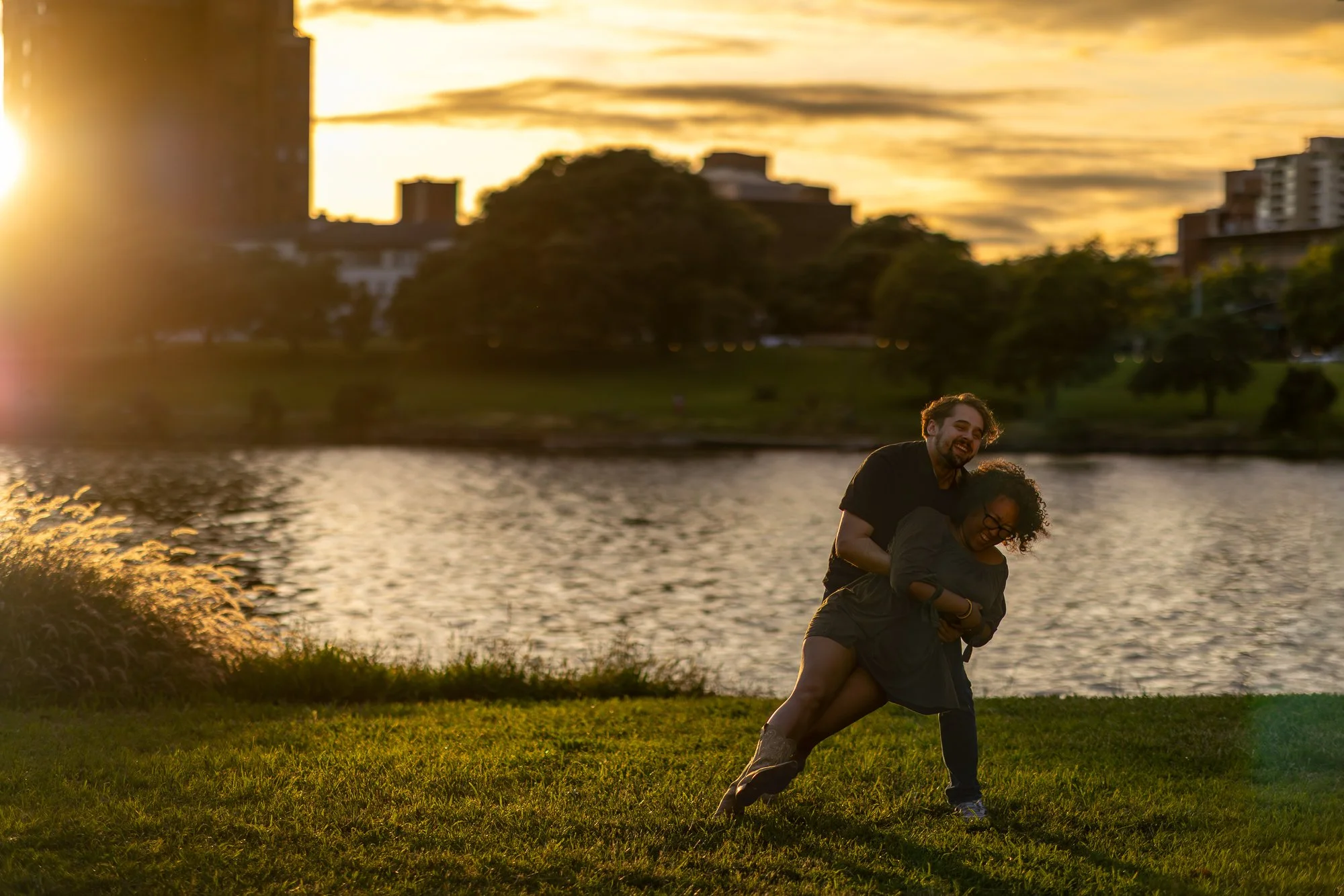 Two people, a man and a woman, are playing around by a lake during sunset, with the man giving the woman a piggyback ride. The background shows trees and buildings silhouetted against a cloudy sky.