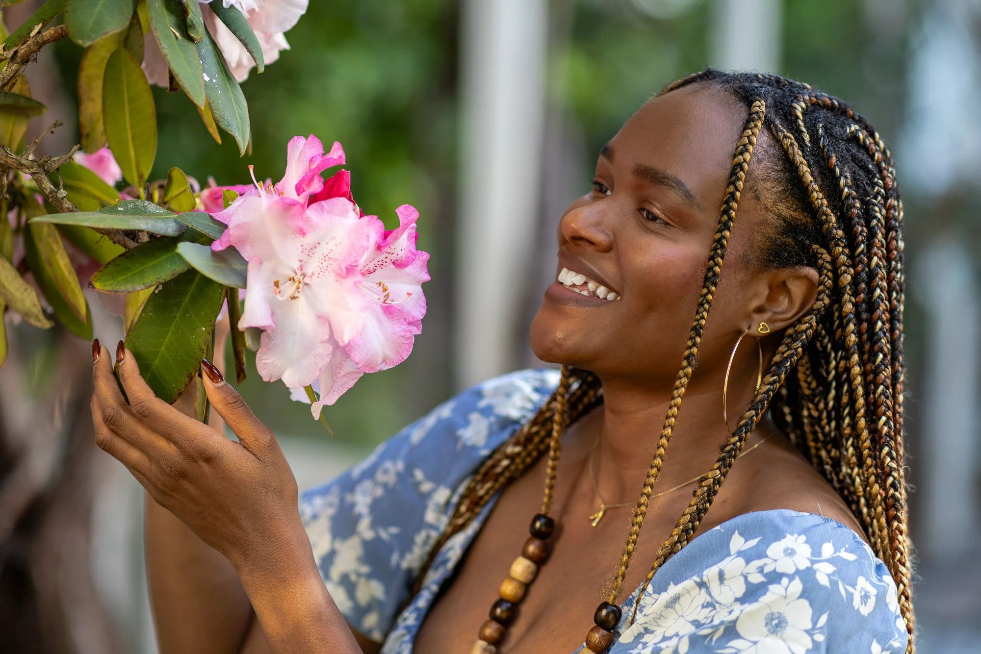 A woman with braided hair, wearing earrings and a necklace, smiling and touching pink and white flowers on a bush.