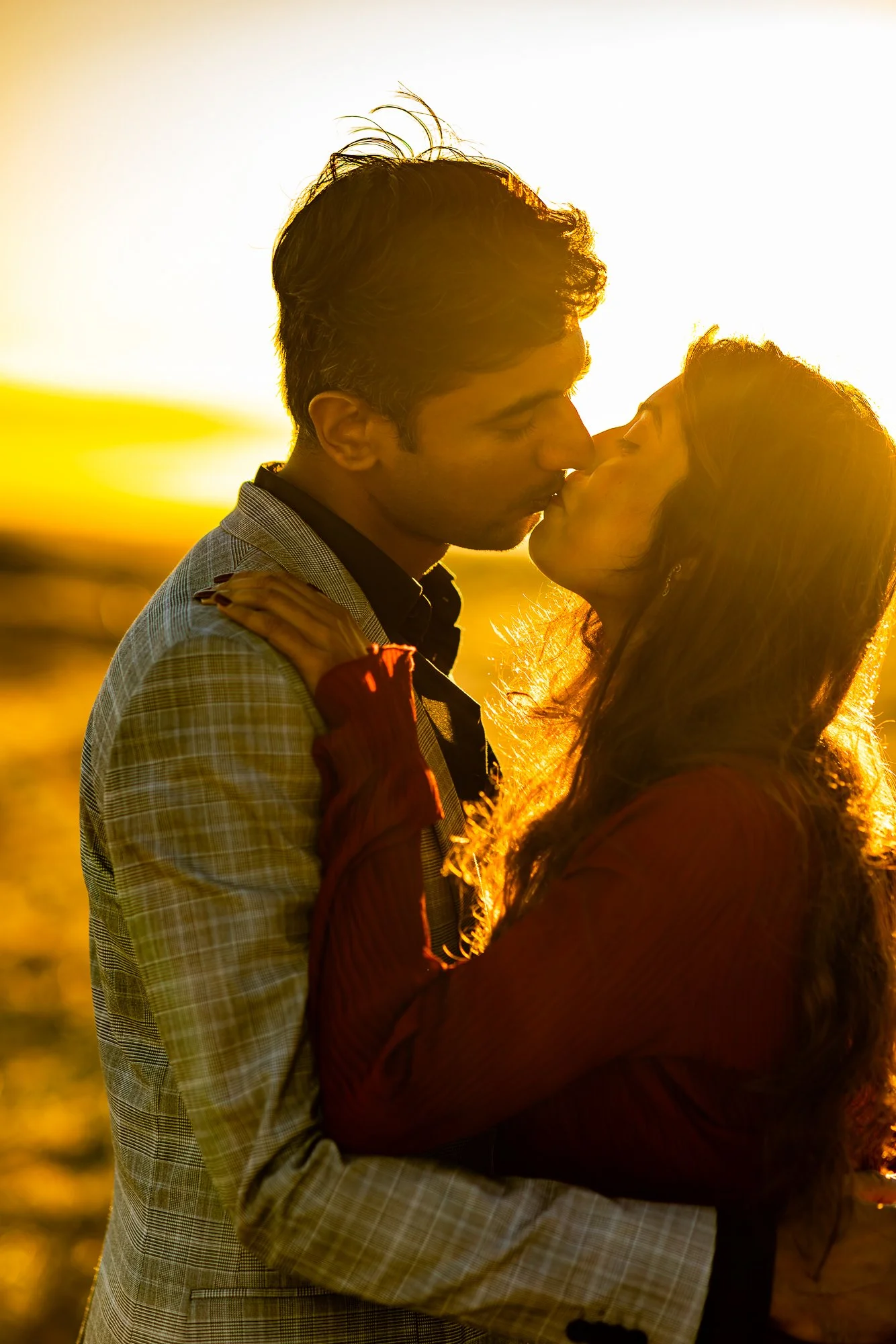 A couple sharing a kiss at sunset, with the sunlight backlighting them and highlighting their profile and hair, in an outdoor setting.
