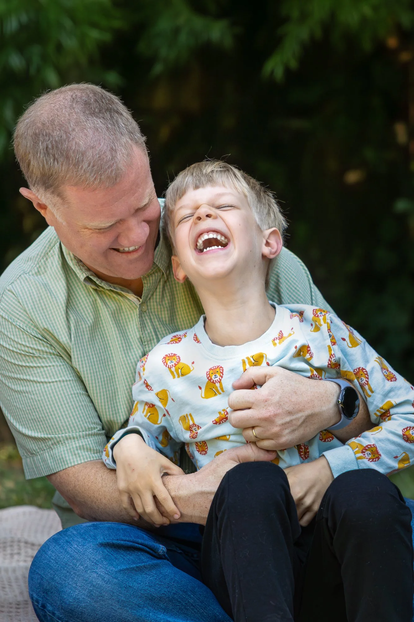 An older man and a young boy are sitting outdoors, laughing and embracing each other. The boy is wearing a white shirt with a lion print, and the man is wearing a green striped shirt. The background features green foliage.