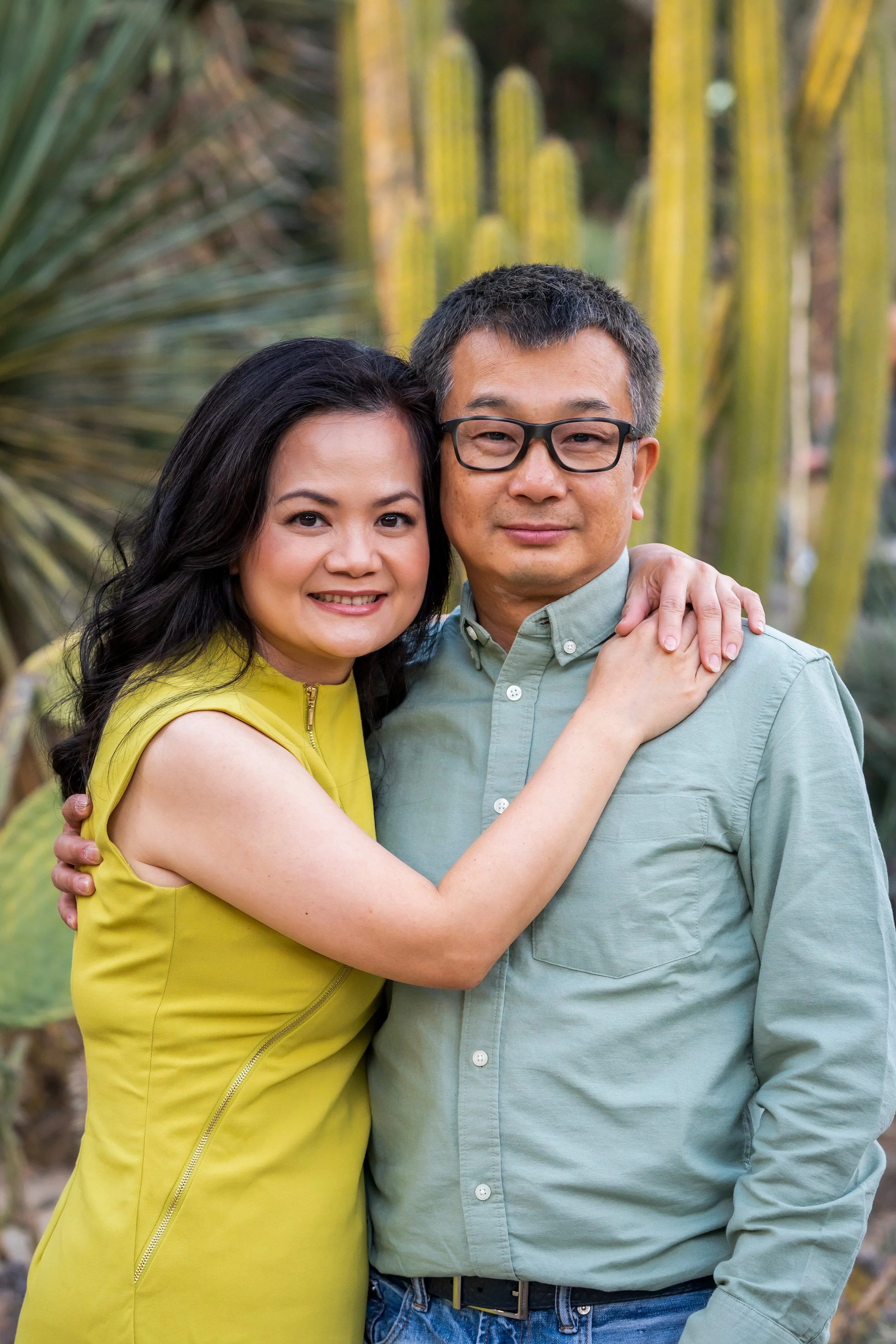 A smiling woman with dark hair in a yellow sleeveless dress hugging a man with glasses, short dark hair, and wearing a light green button-up shirt, standing outdoors with tall green cacti in the background.