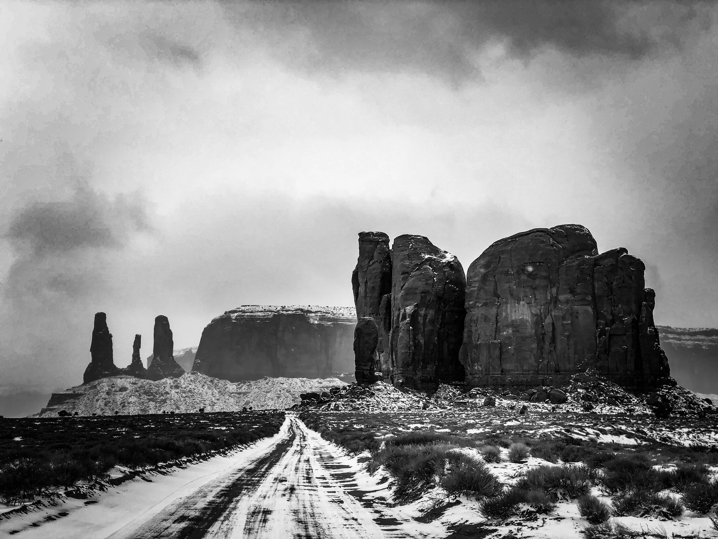 Black and white photo of a rugged desert landscape with towering rock formations, a snow-covered dirt road in the foreground, and dark clouds overhead.