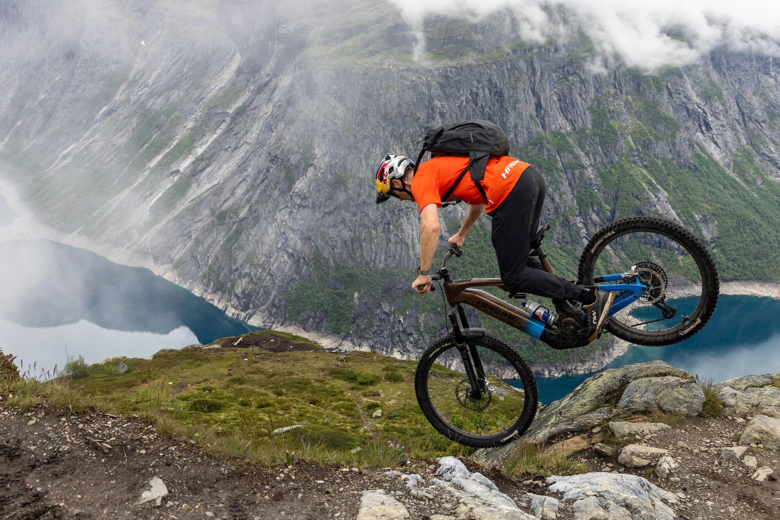 Mountain biker wearing a helmet and orange shirt riding down a rocky trail with a scenic fjord and steep cliffs in the background.