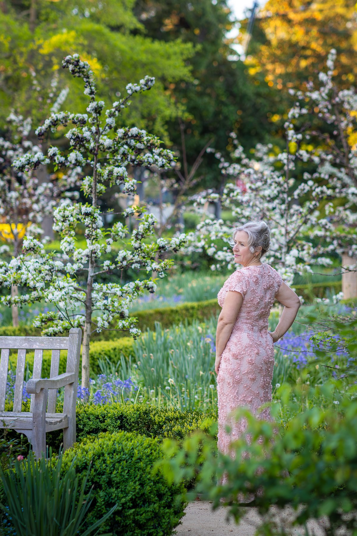 Woman in pink floral dress standing in spring garden looking back over shoulder, apple blossom trees and blue flowers behind her, Gamble Garden Palo Alto