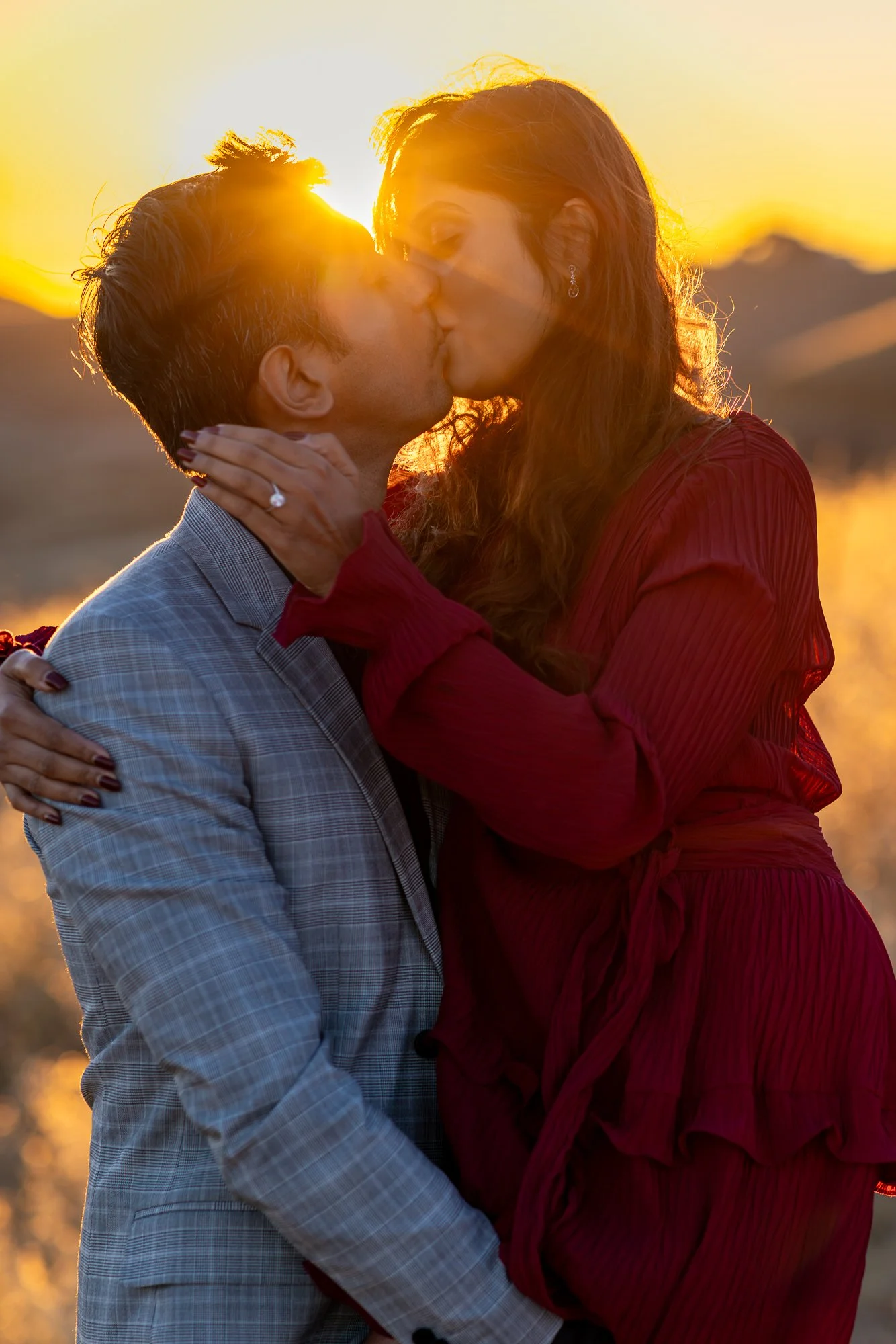 A couple kissing outdoors at sunset, with the sun setting behind them and mountains in the background. The woman is wearing a red dress and the man a light suit.