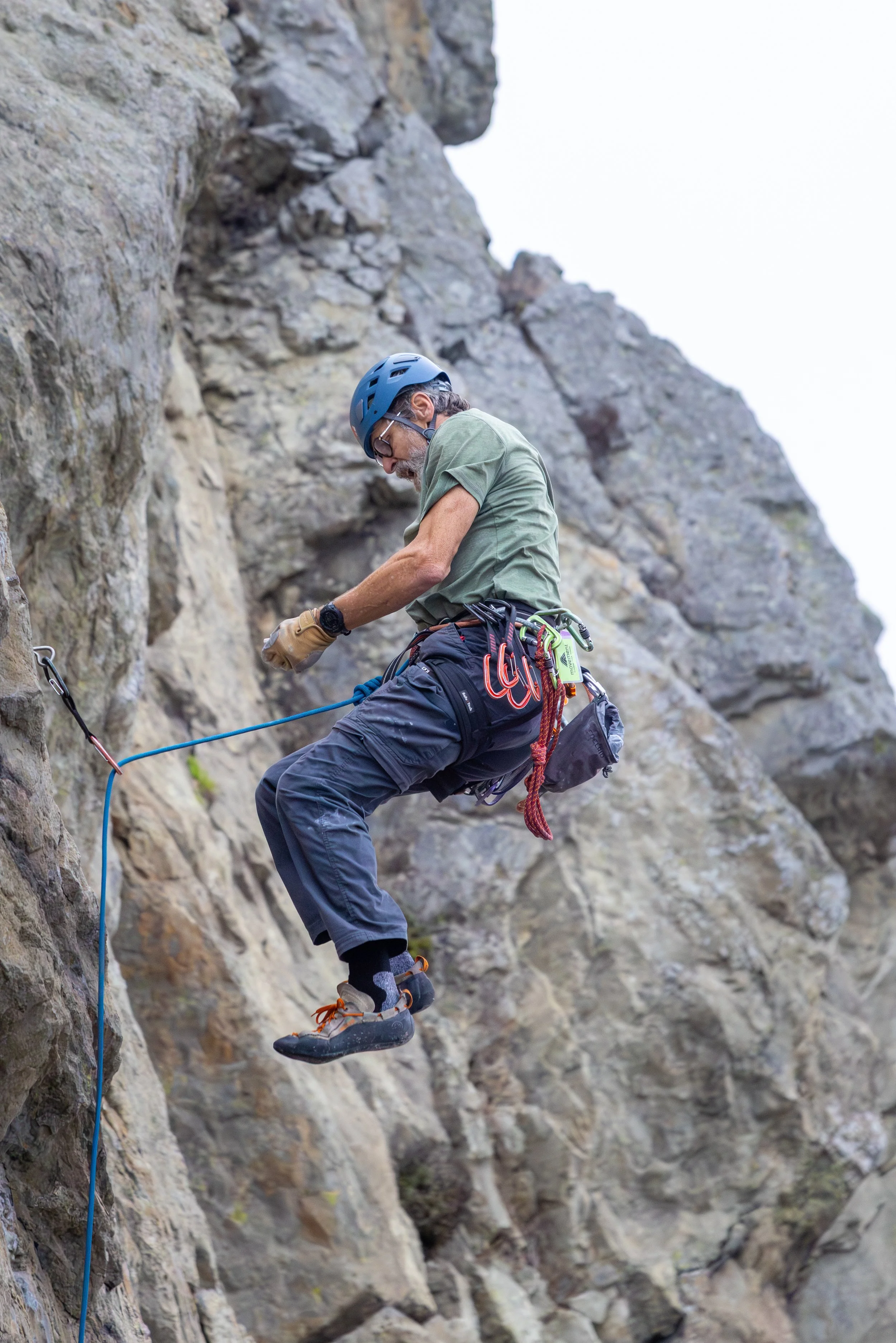 An older man rock climbing on an outdoor rock face, wearing a blue helmet, green t-shirt, black pants, and climbing shoes, with climbing gear attached to his harness.