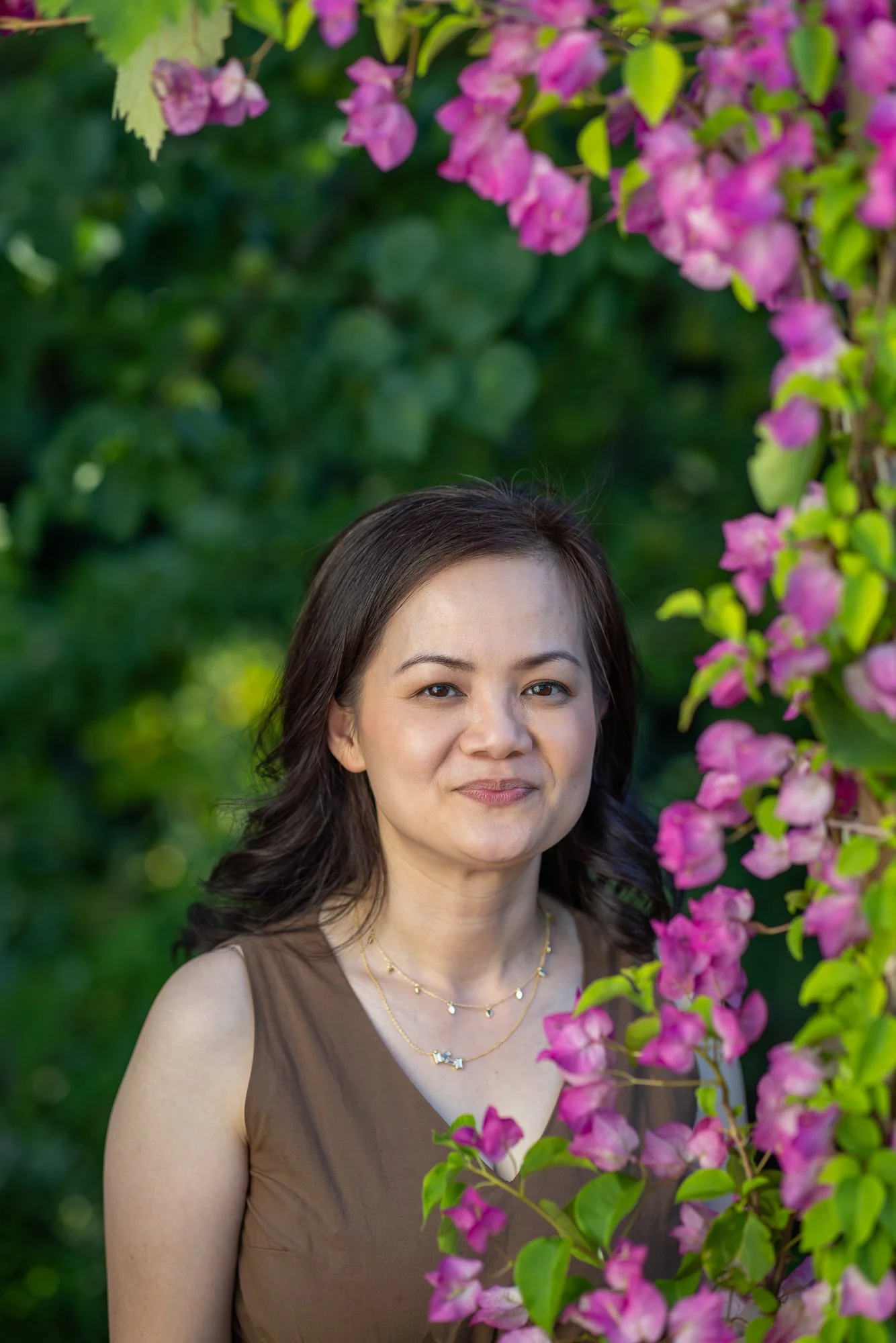 A woman with shoulder-length dark hair and wearing a beaded necklace stands beside pink flowers with green leaves, in an outdoor garden setting.