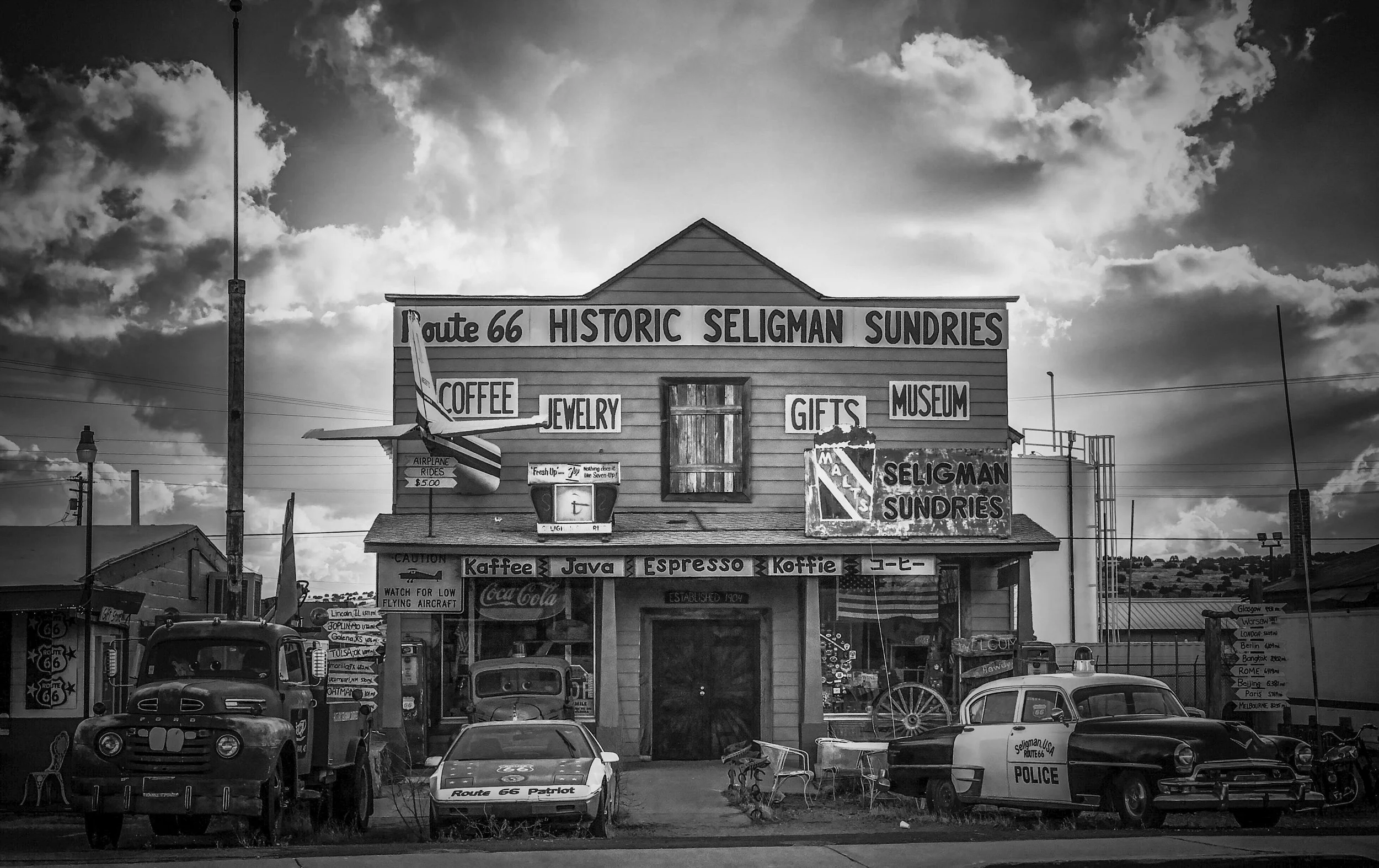 Black and white photo of a vintage roadside store with signs advertising Route 66 Seligman Sundries, coffee, jewelry, gifts, and a museum. Several old cars are parked in front, and the sky is partly cloudy.