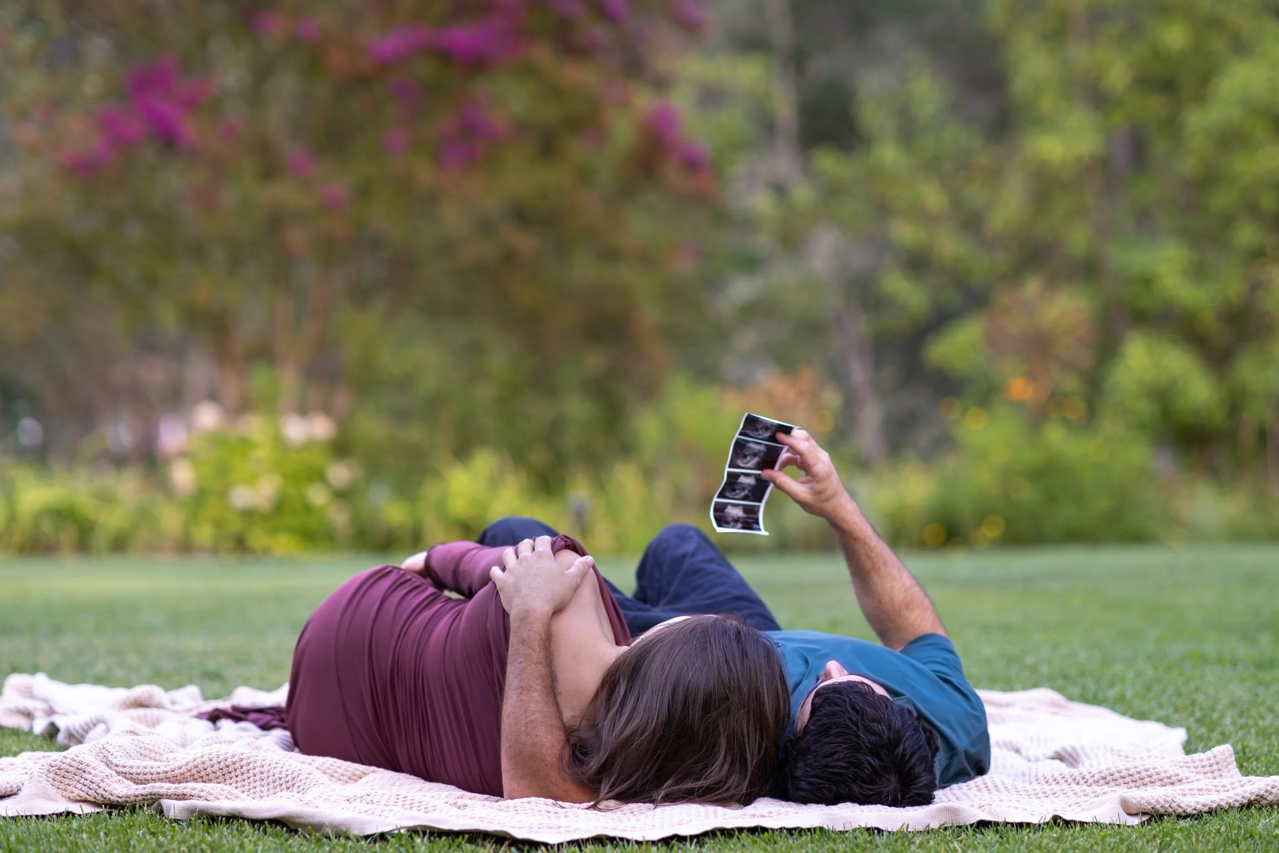 A couple lying on a blanket in a park, taking turns holding and looking at ultrasound photos, with trees and pink flowers in the background.