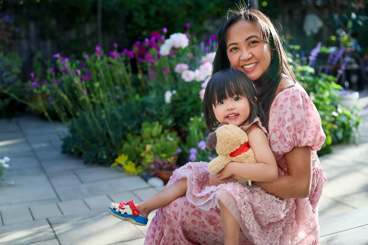 A smiling woman and young girl sitting outdoors in a garden, the girl holding a teddy bear, surrounded by colorful flowers.