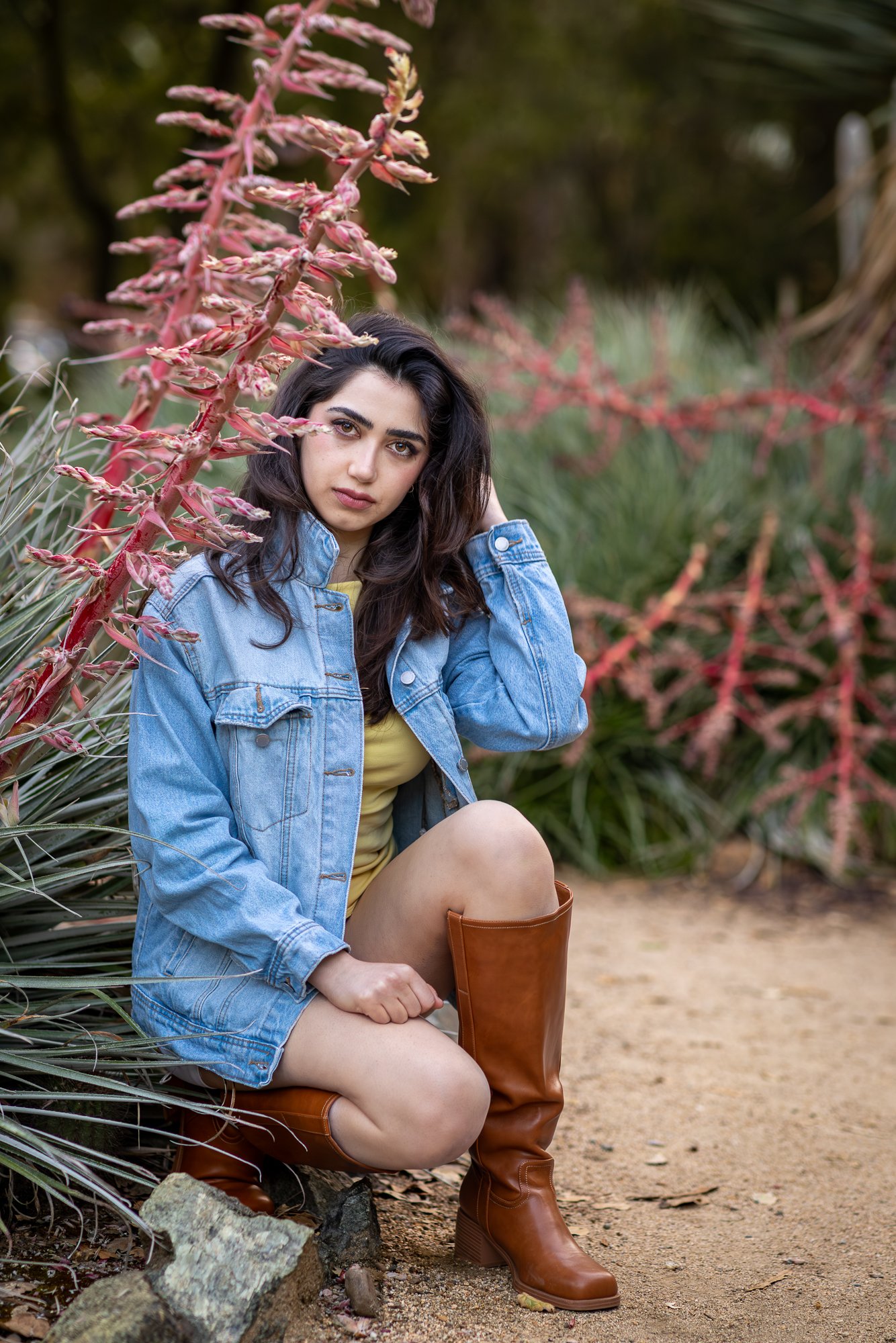 A woman sitting outdoors in a natural setting with plants and trees in the background. She is wearing a denim jacket, yellow dress, and brown knee-high boots, with one knee bent and her hand resting on her head, looking at the camera.
