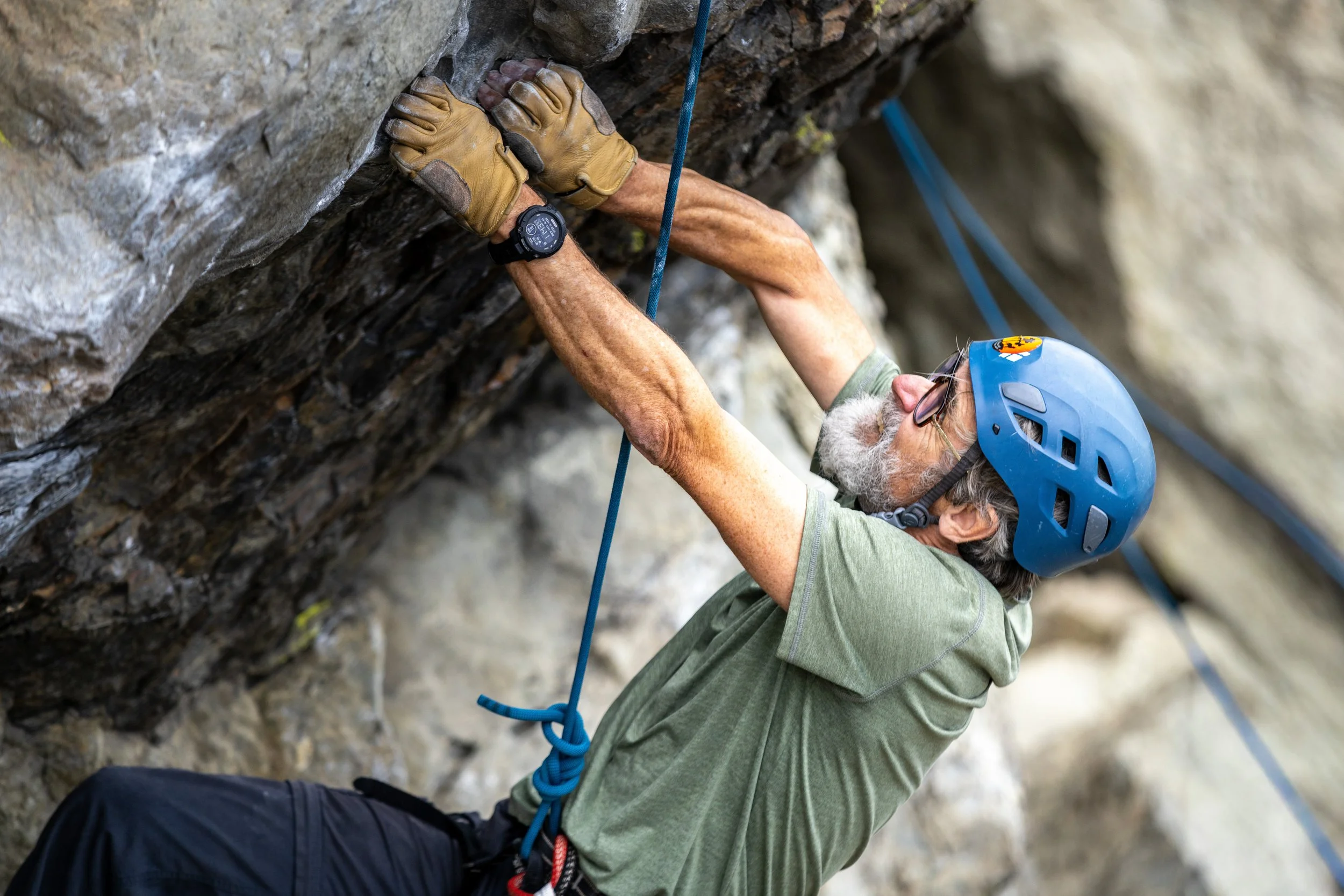 An older man rock climbing on an indoor or outdoor rock wall, wearing a blue helmet, glasses, a green shirt, and a watch, with climbing gloves, holding onto the rock with both hands.