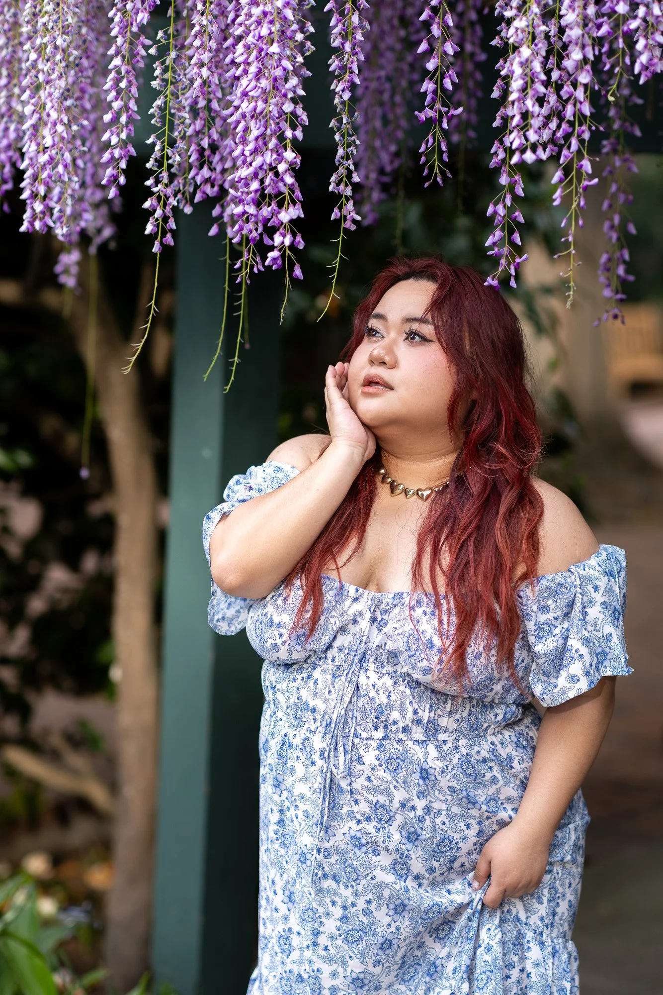 Woman in blue floral off-shoulder dress with hand near jaw looking up at wisteria cascading from pergola, natural light portrait Gamble Garden Palo Alto
