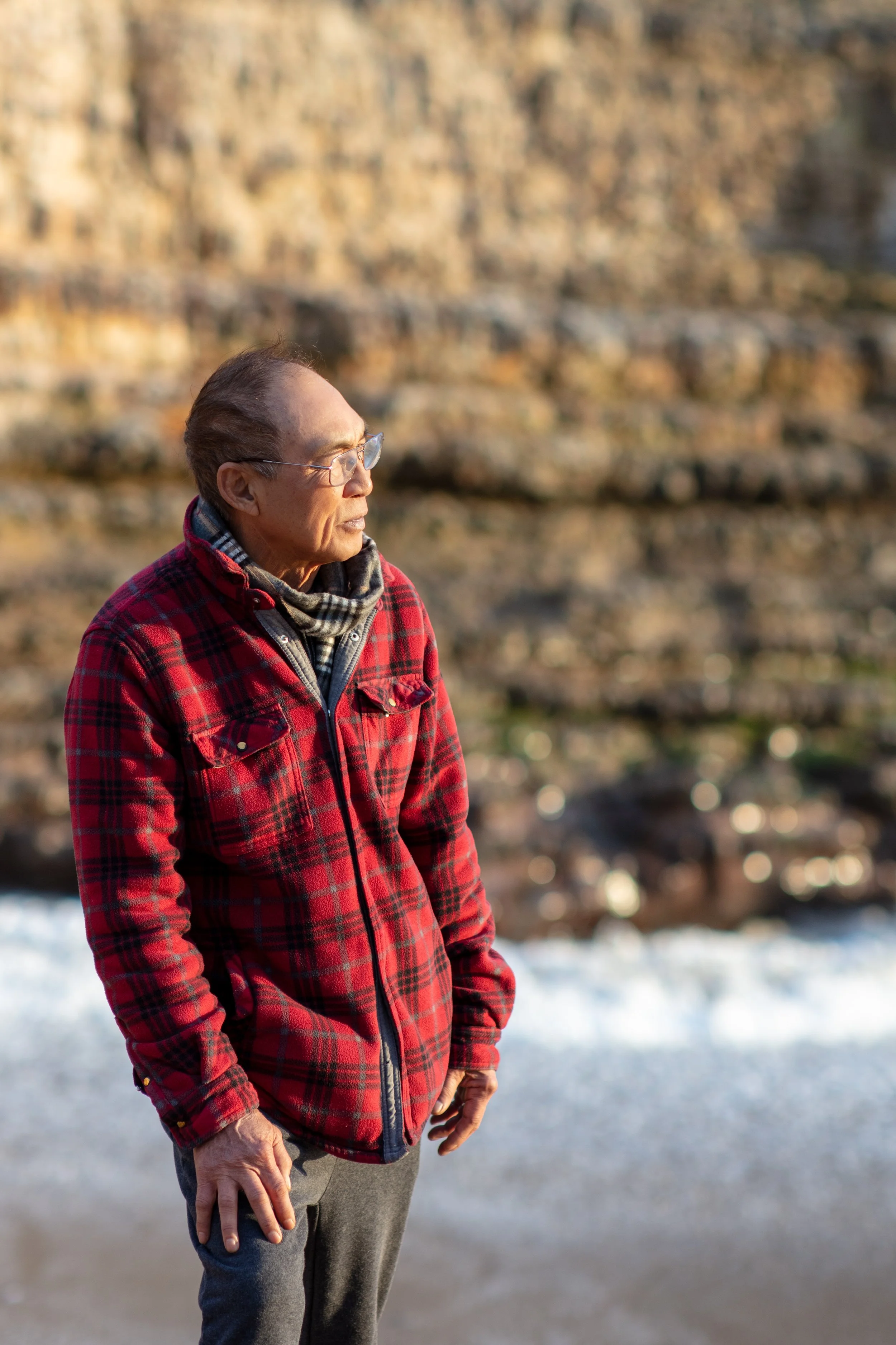 An elderly man stands outdoors, wearing a red plaid jacket, gray pants, glasses, and a scarf, looking towards the horizon in front of a rocky cliff with water below.