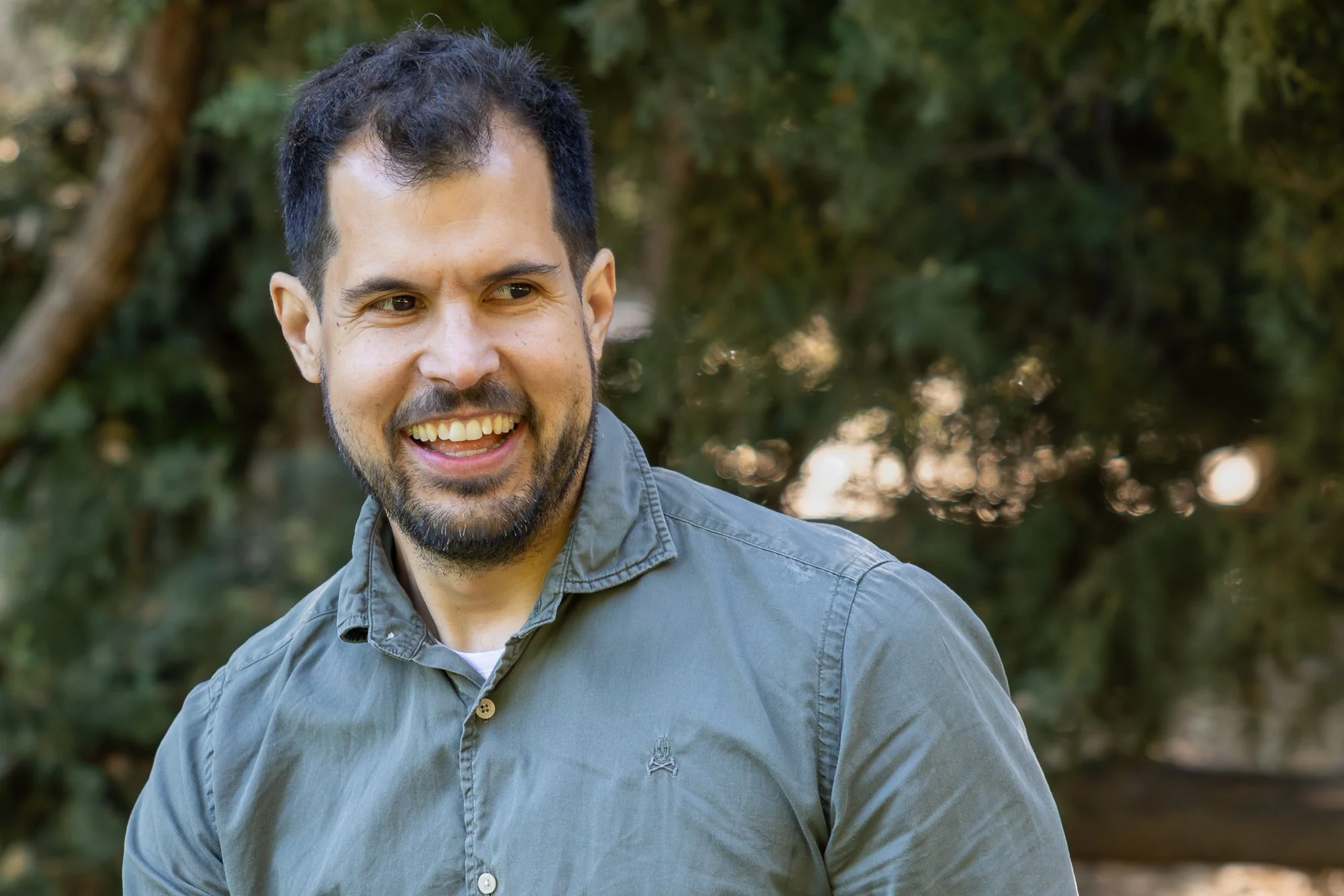 A smiling man with dark hair, beard, and mustache wearing a gray button-up shirt outdoors.