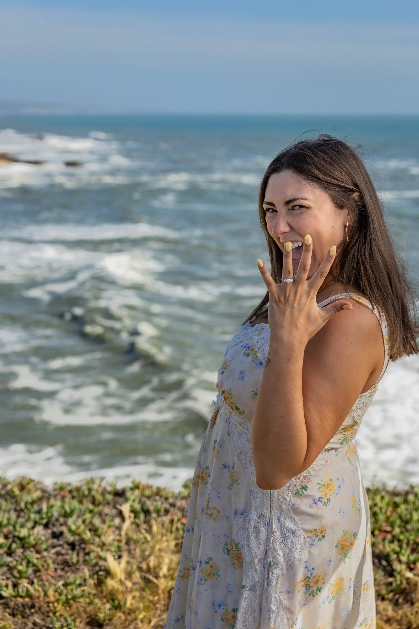 A woman in a floral dress with yellow nails and gold hoop earrings stands near the ocean, smiling and covering her mouth with her hand, with waves and a blue sky in the background.