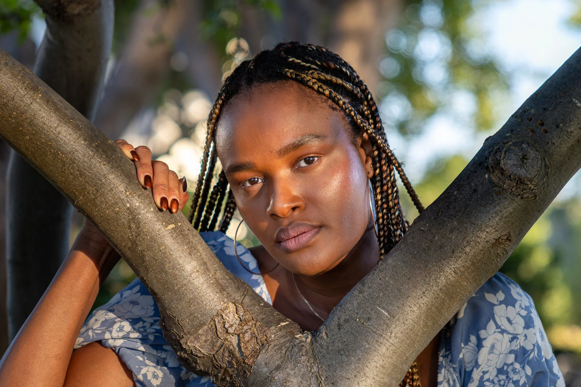 A young woman with braided hair and hoop earrings poses behind the branches of a tree, looking directly at the camera with a serene expression.