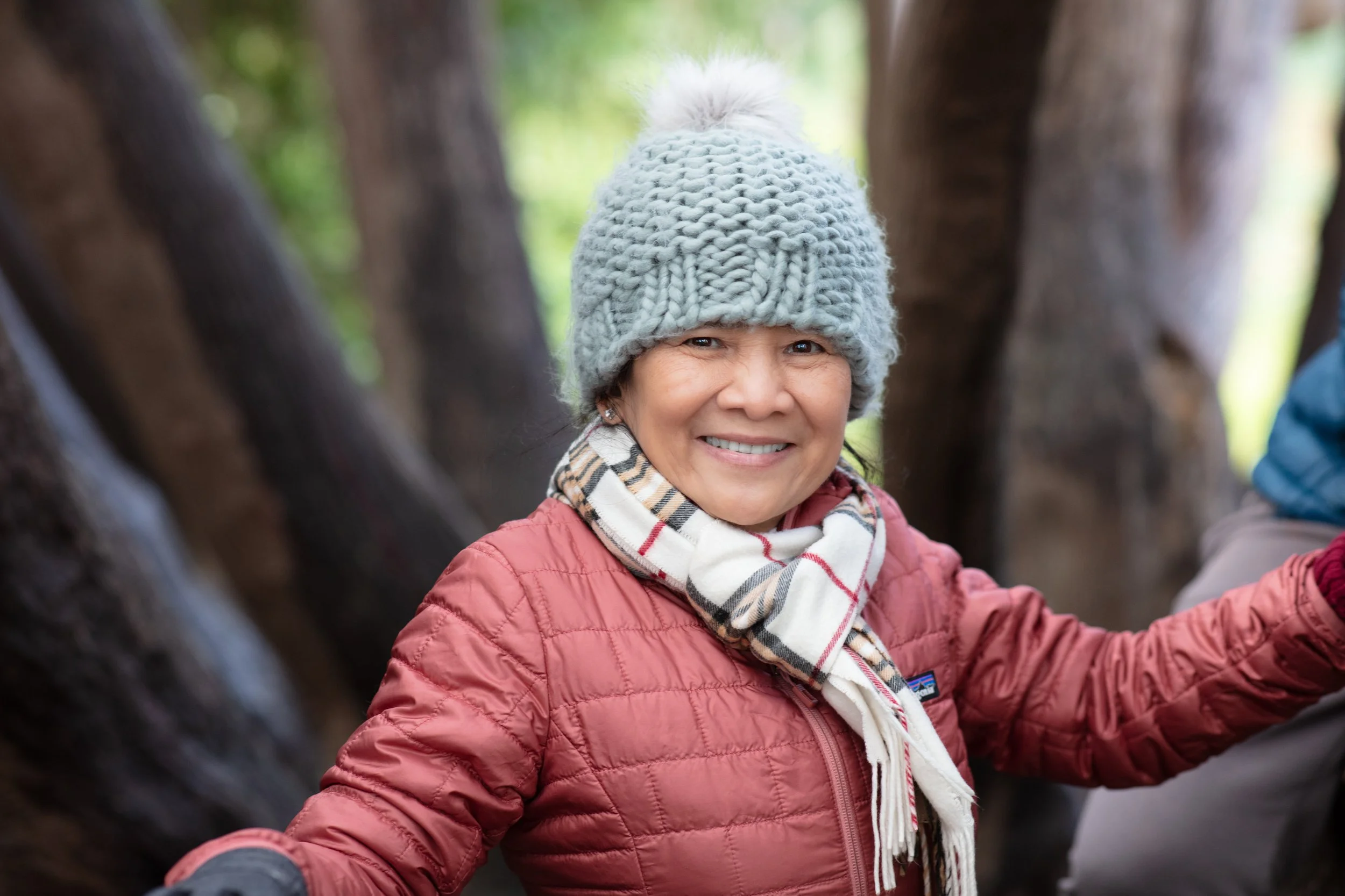 A smiling woman wearing a gray knit beanie, a plaid scarf, and a red quilted jacket outdoors with trees in the background.