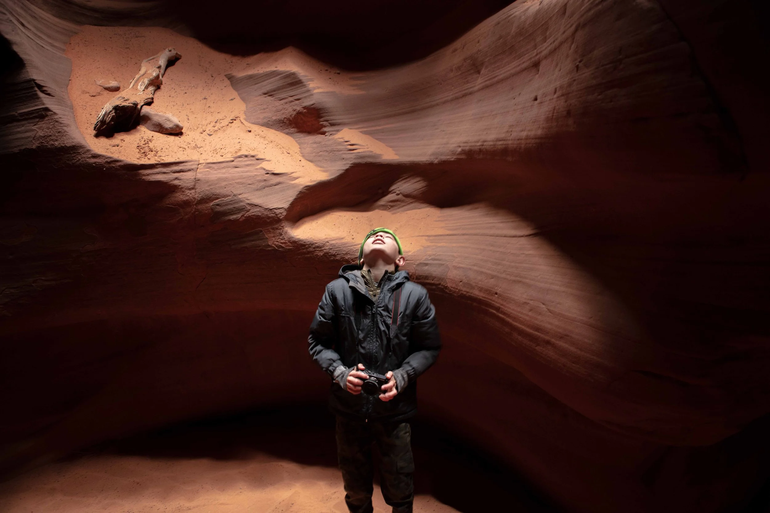 A person standing inside a narrow slot canyon with smooth, layered reddish-brown rock formations, holding a camera and looking up.