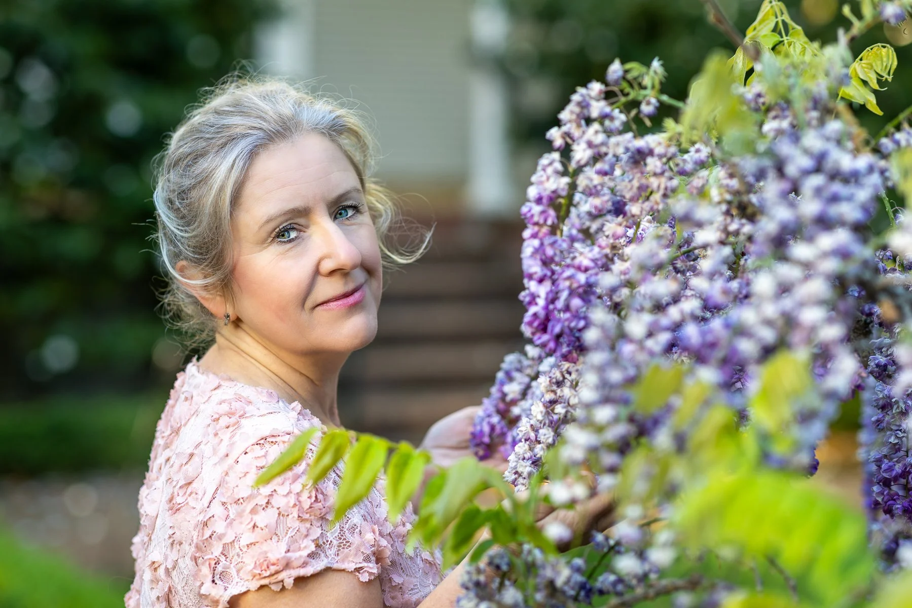 Woman in blush pink dress holding wisteria blooms and looking directly at camera, morning light portrait at Elizabeth Gamble Garden
