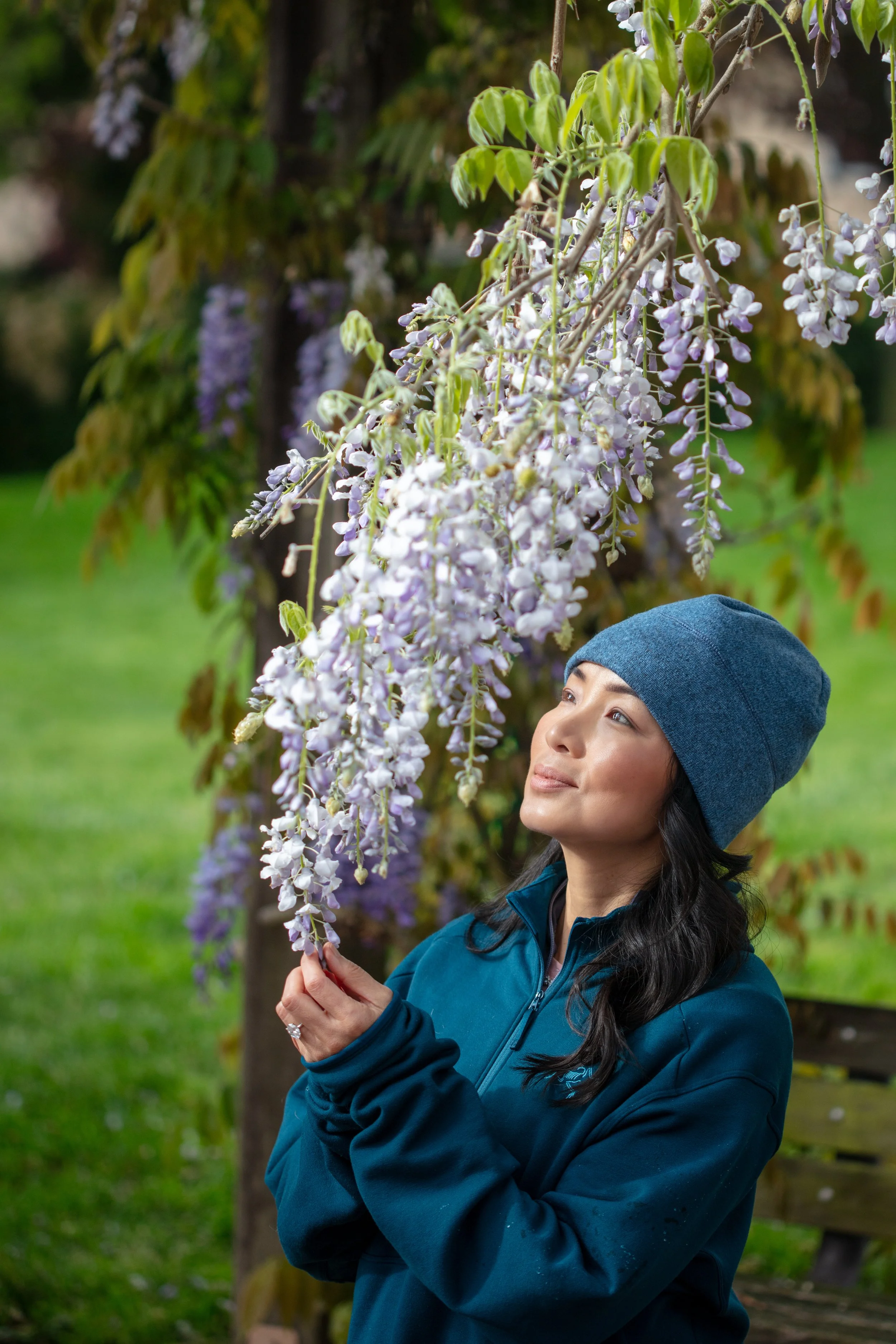 A woman wearing a blue beanie and teal jacket being near a flowering wisteria tree with purple hanging blossoms in a park.