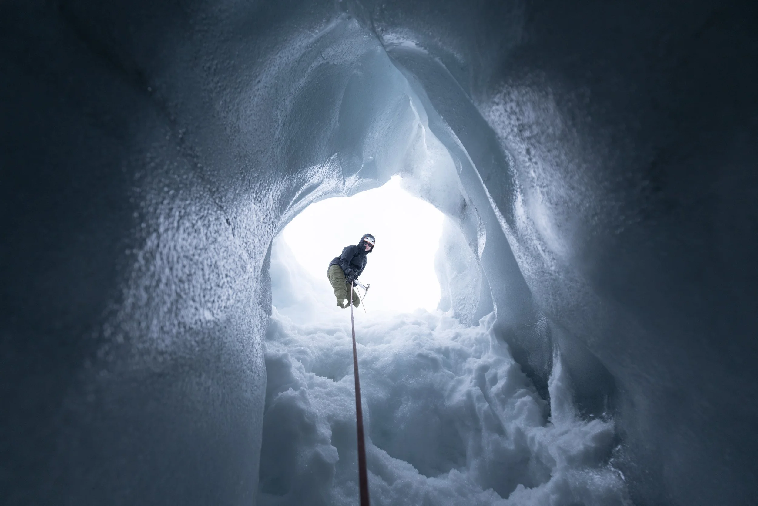 A person inside an ice cave, seen from below, holding an ice axe, with bright light outside the icy opening.