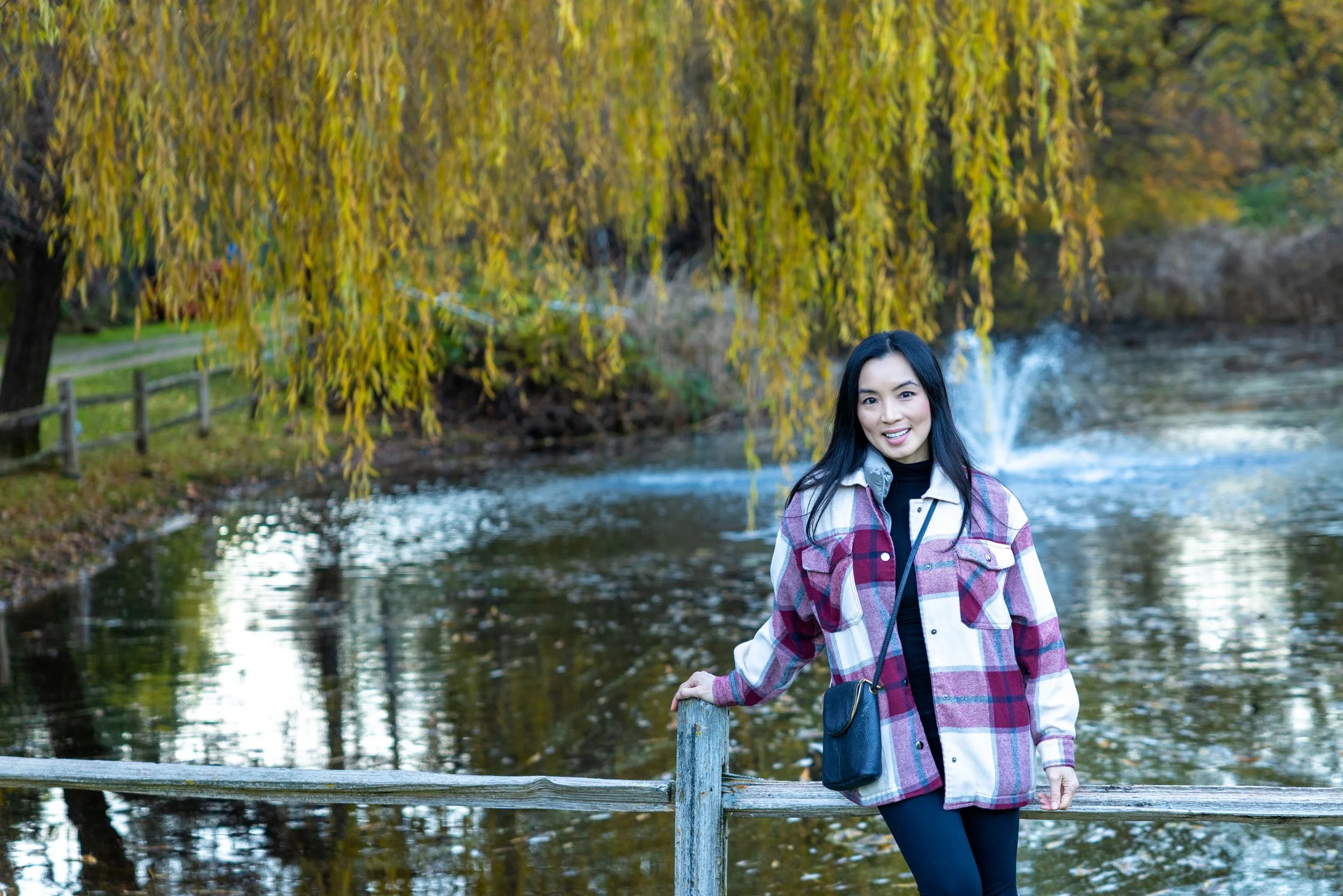 A woman smiling and standing by a river with autumn-colored trees in the background.