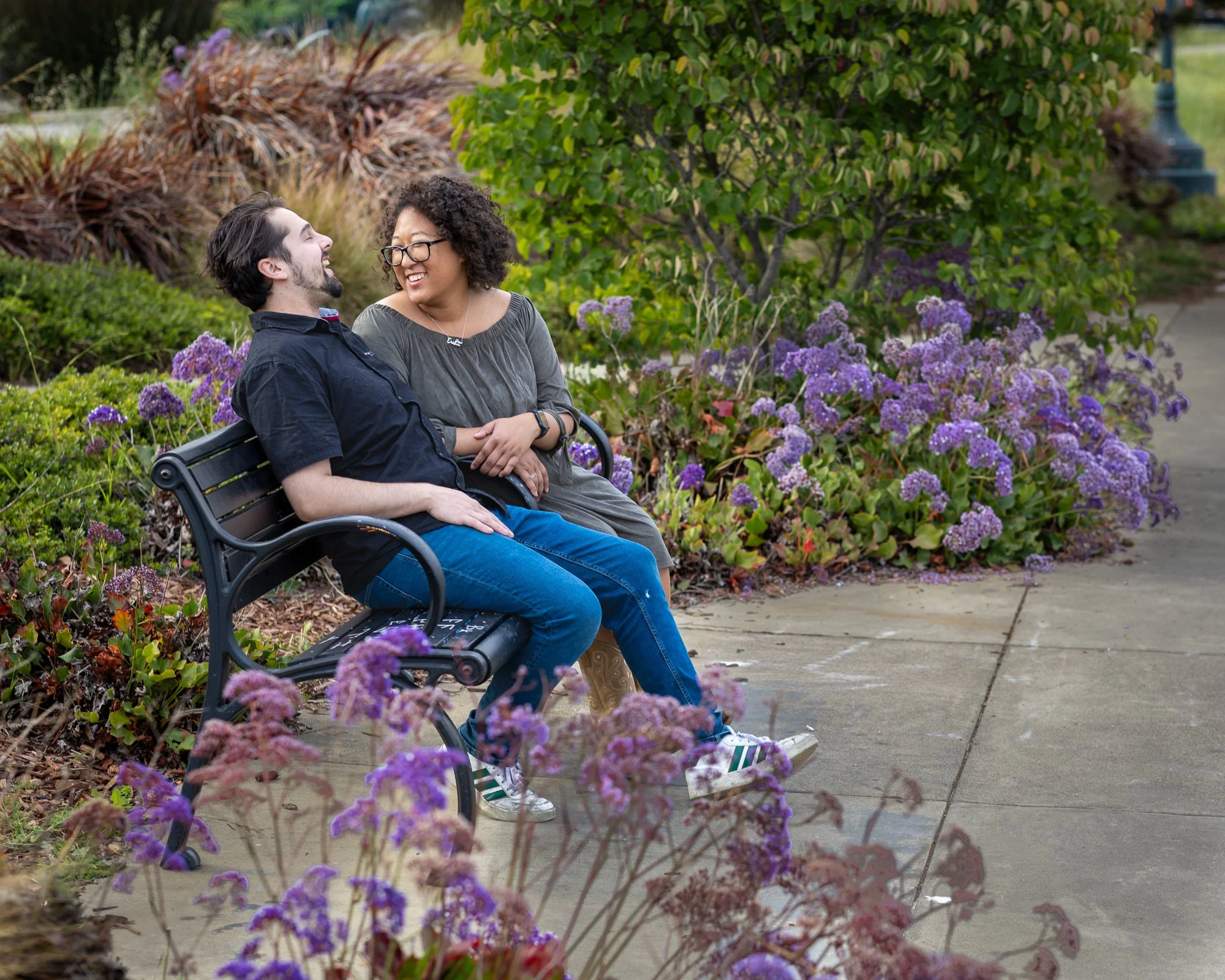 A man and woman sitting on a park bench, smiling at each other, surrounded by purple flowers and green bushes.