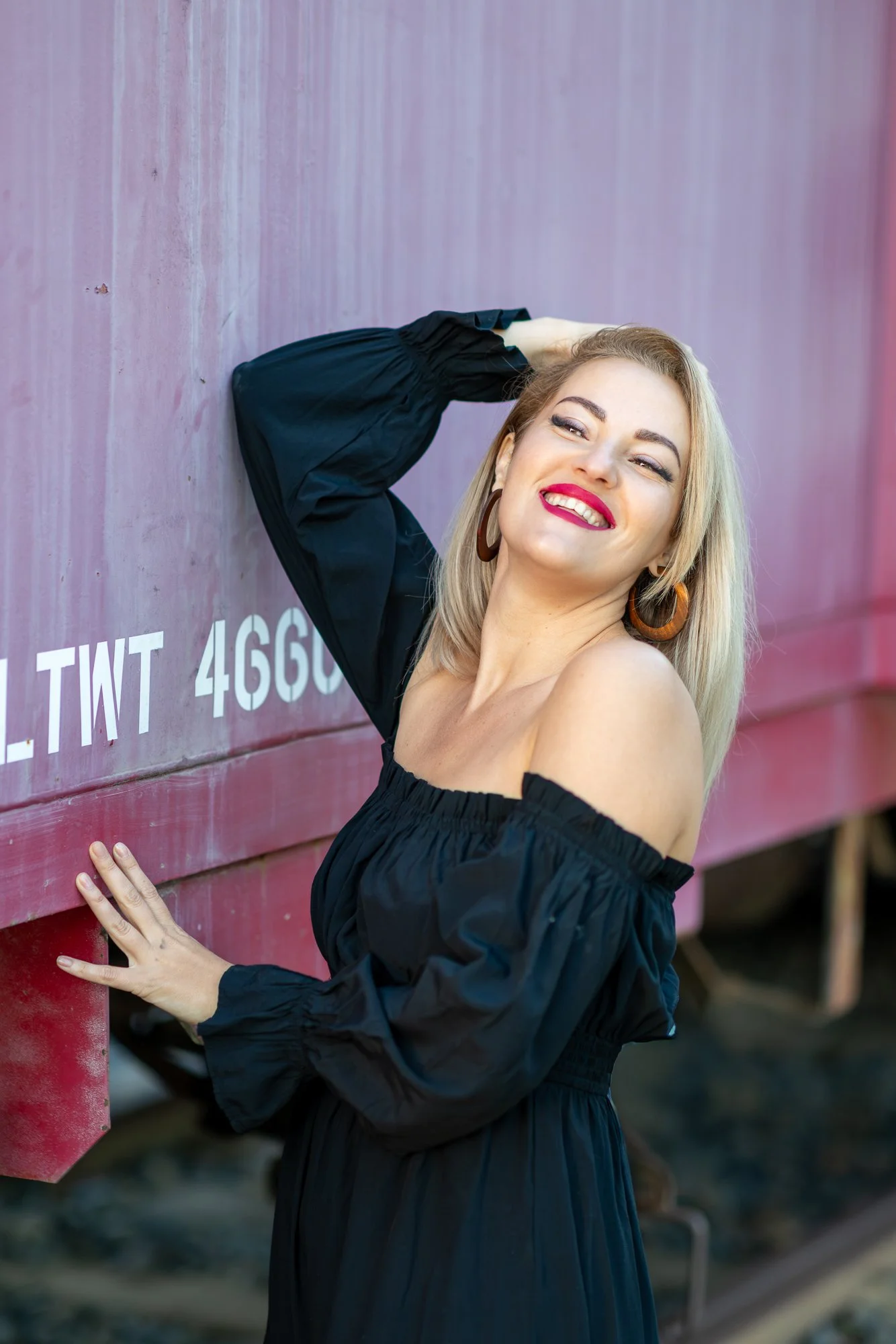 A woman with blonde hair and large hoop earrings smiling and leaning against a pink train car, wearing an off-shoulder black dress.
