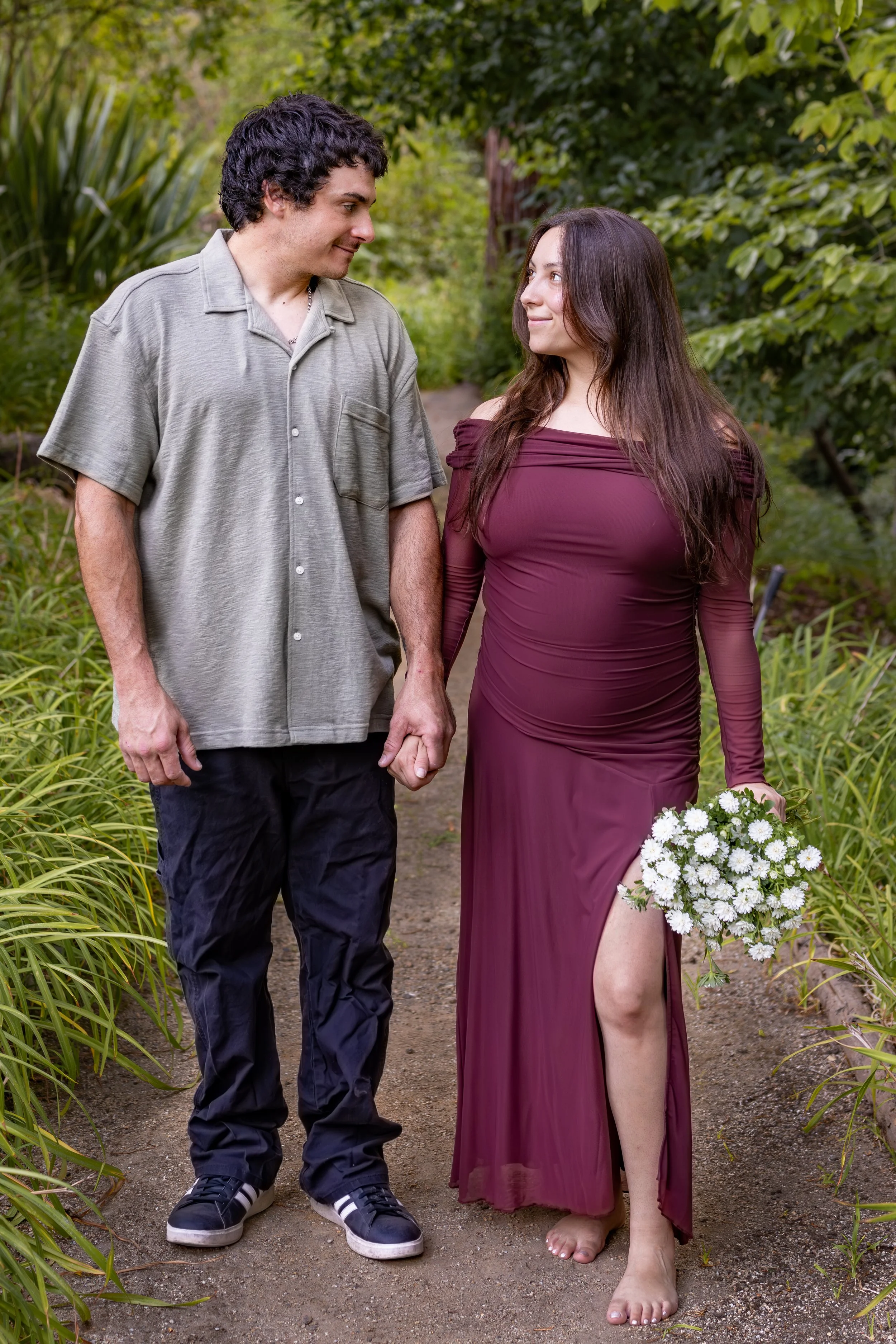 A man and woman holding hands and smiling at each other on a garden path, with greenery and plants around them. The woman wears a purple dress and holds a bouquet of white flowers.