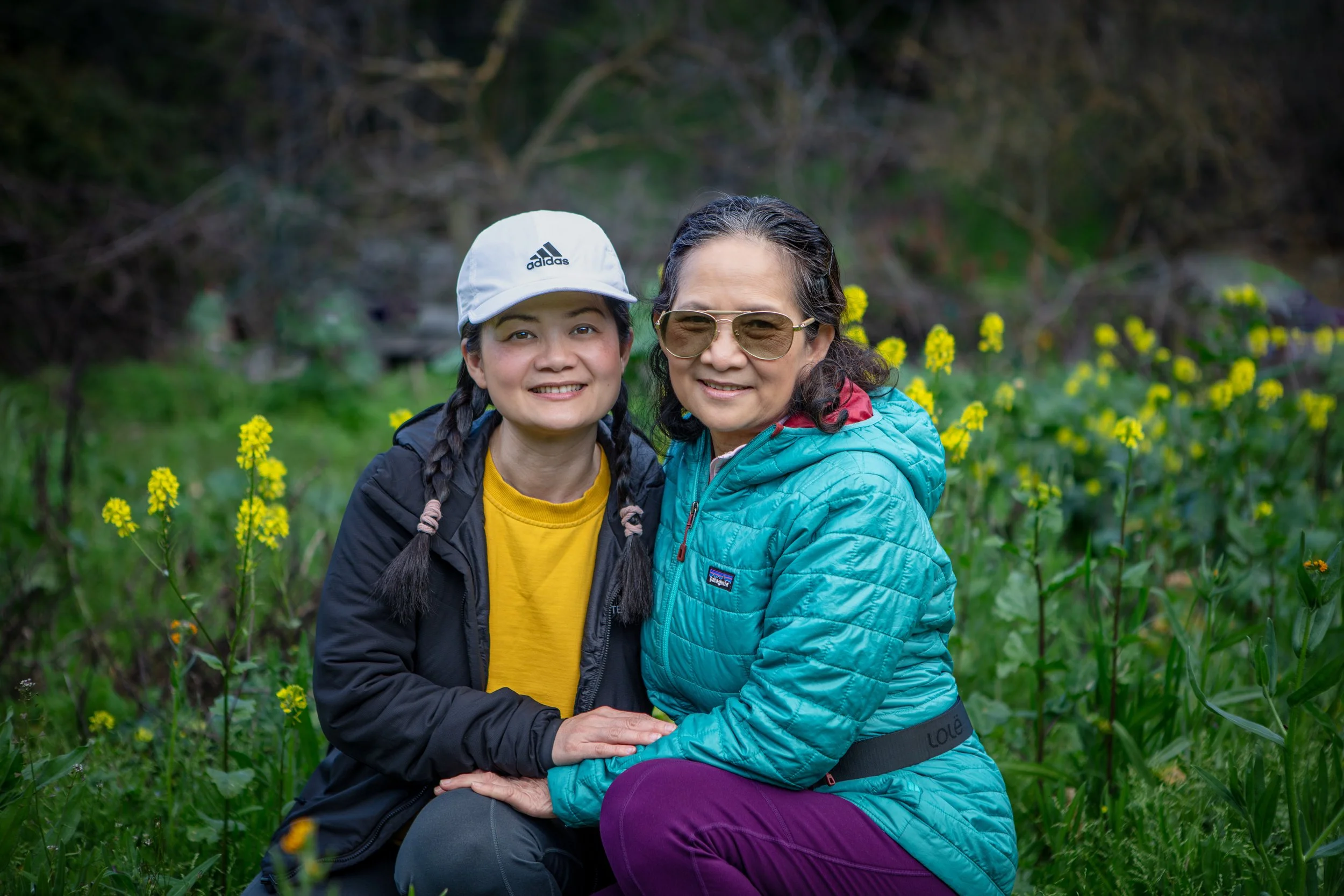 Two women sitting in a field of yellow flowers, smiling, with greenery and trees in the background.